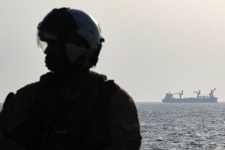 A commercial vessel is seen in the background as a US Black Hawk helicopter crew member stands on the deck of Britain's RFA Cardigan Bay landing ship in the Gulf waters off Bahrain during the International Maritime Exercise (IMX), on November 5, 2019. - IMX is a joint military exercise involving assets and personnel from more than 50 partner nations and seven international organisations. The US has pushed for the creation of a US-led operation dubbed the International Maritime Security Construct to safeguard trade and the flow of oil through the Strait of Hormuz. It has so far been joined by Australia, Britain and the United Arab Emirates, as well as Bahrain, the Gulf island state which is home to the US Fifth Fleet.