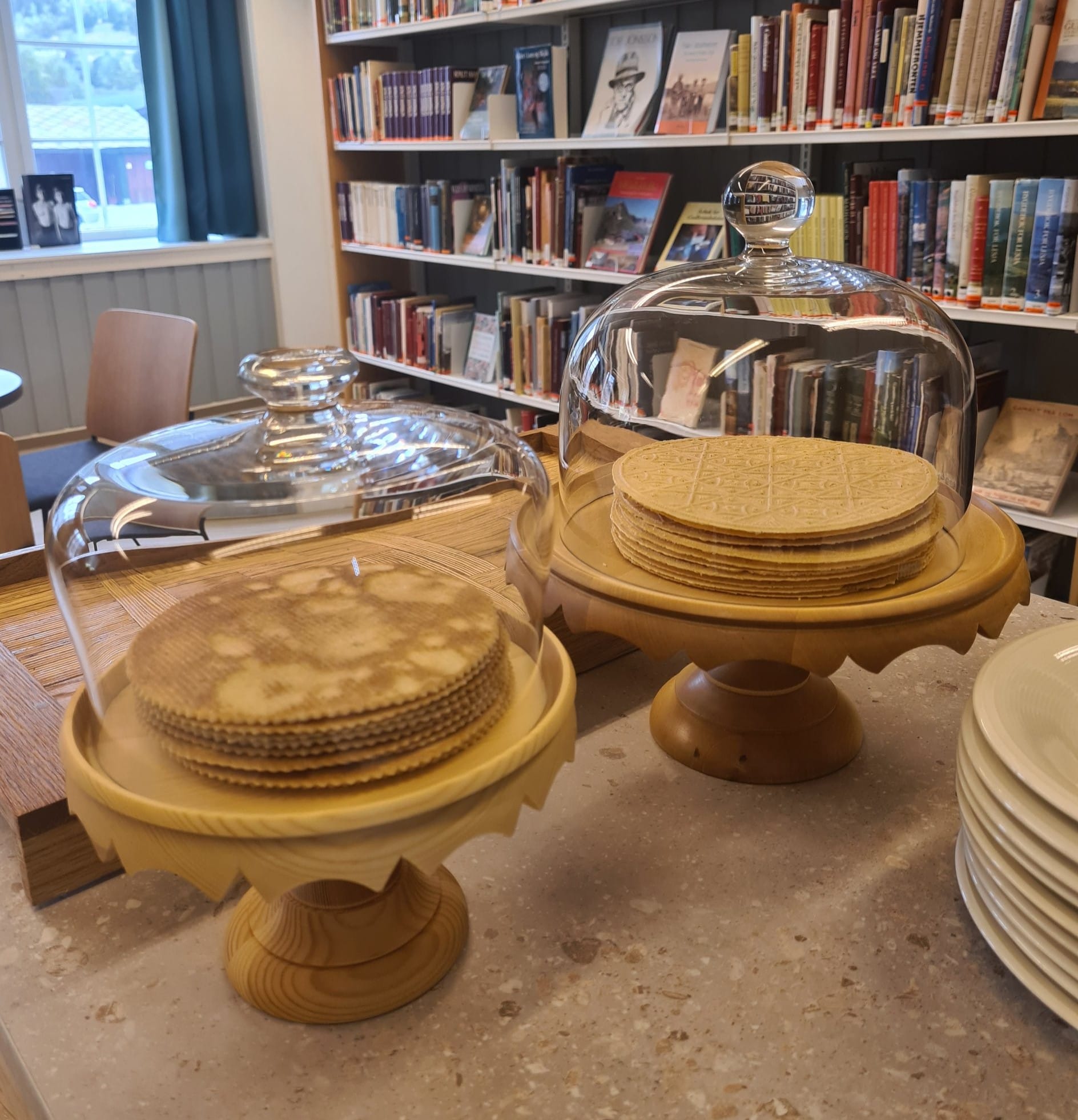 Stacks of waffles under glass domes on wooden cake stands in a room lined with bookshelves.