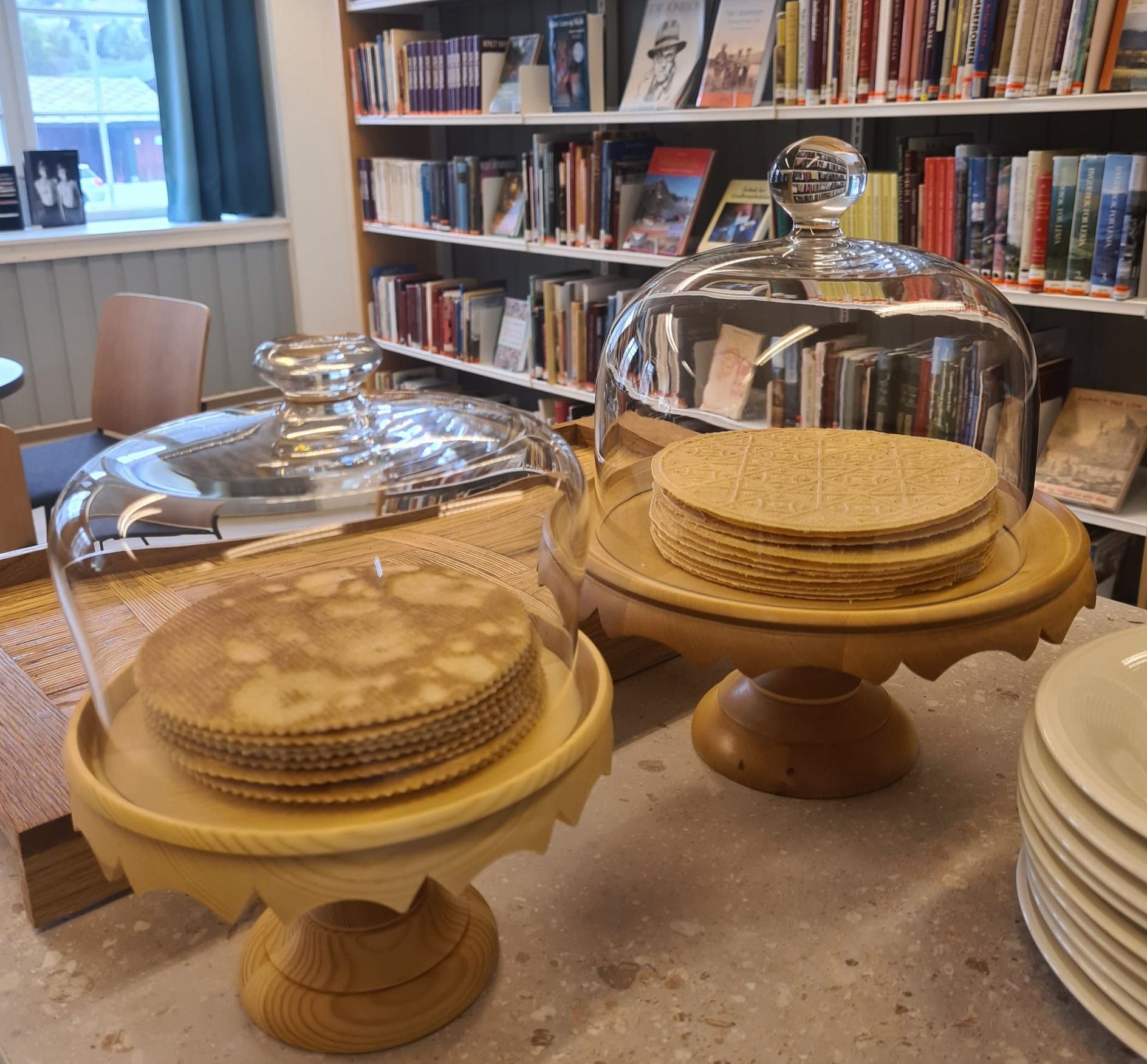 Stacks of waffles under glass domes on wooden cake stands in a room lined with bookshelves.