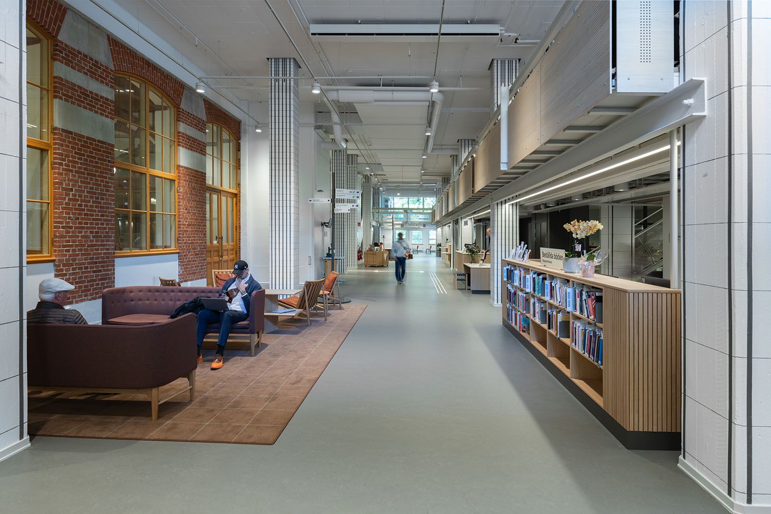 Modern corridor with a lounge area by brick windows on left, and a long wooden book counter on right. People are visible.