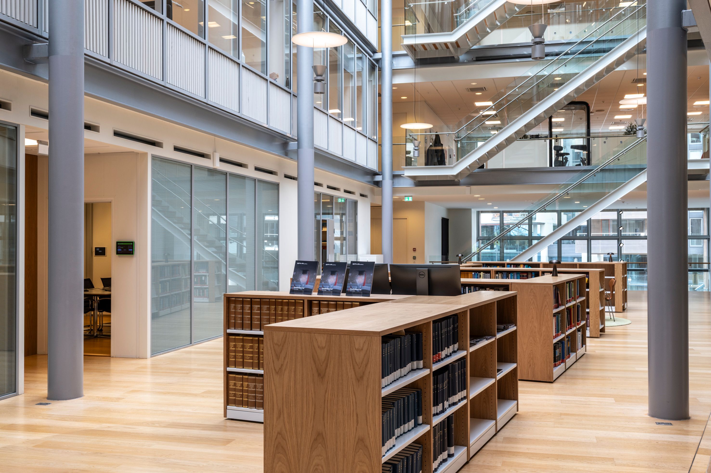 A modern, multi-story library with light wood floors, wooden bookshelves, computers, and a prominent glass staircase.