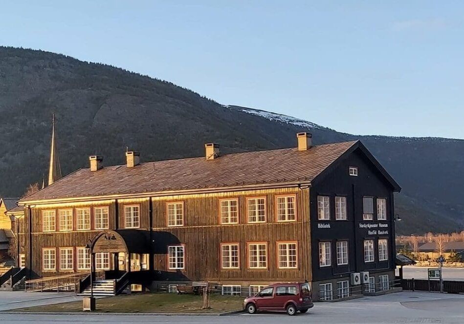 A large dark wooden building with many windows, housing a library and museum, stands before mountains with patches of snow under a clear blue sky.