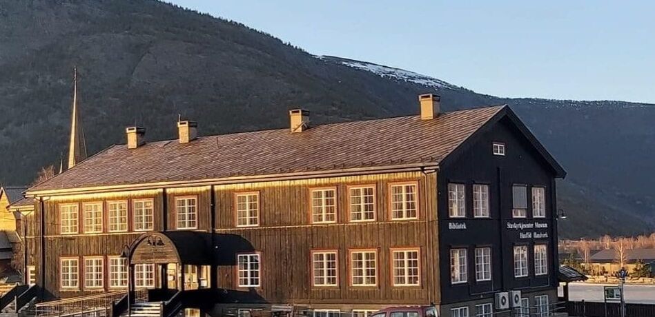 A large dark wooden building with many windows, housing a library and museum, stands before mountains with patches of snow under a clear blue sky.