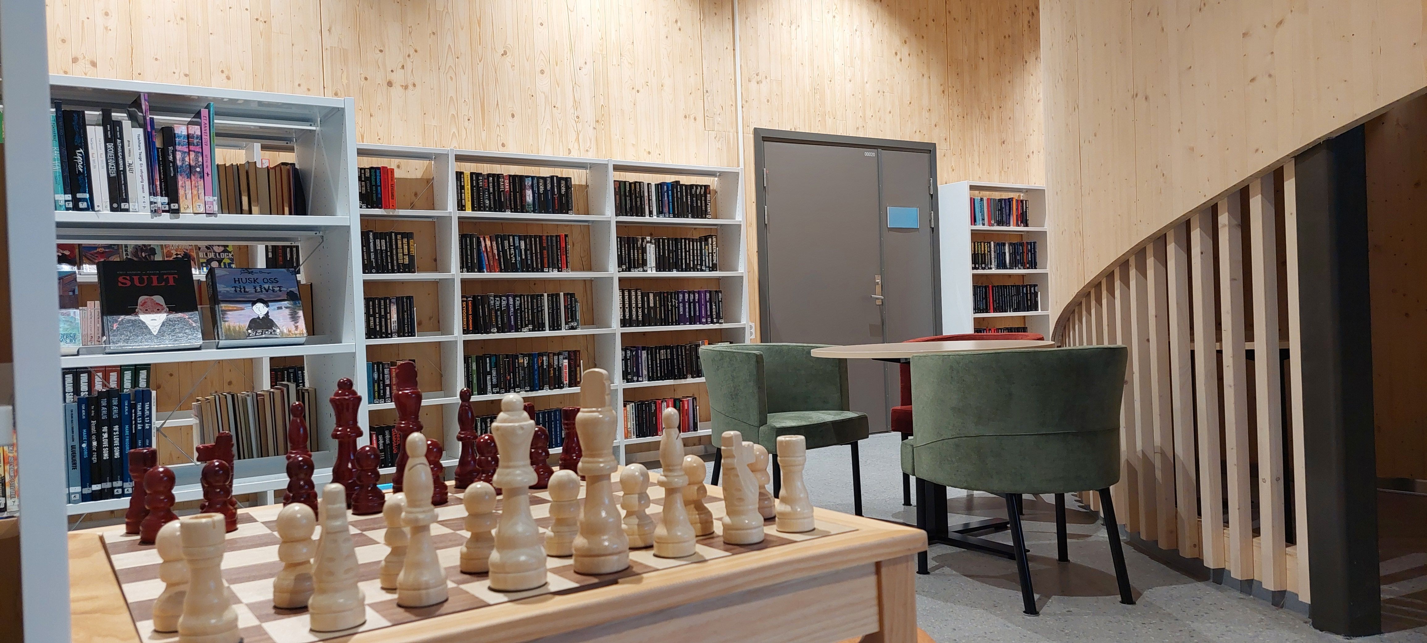 A lounge or library space featuring a large wooden chess set, white bookshelves, green chairs, and wood-paneled walls.