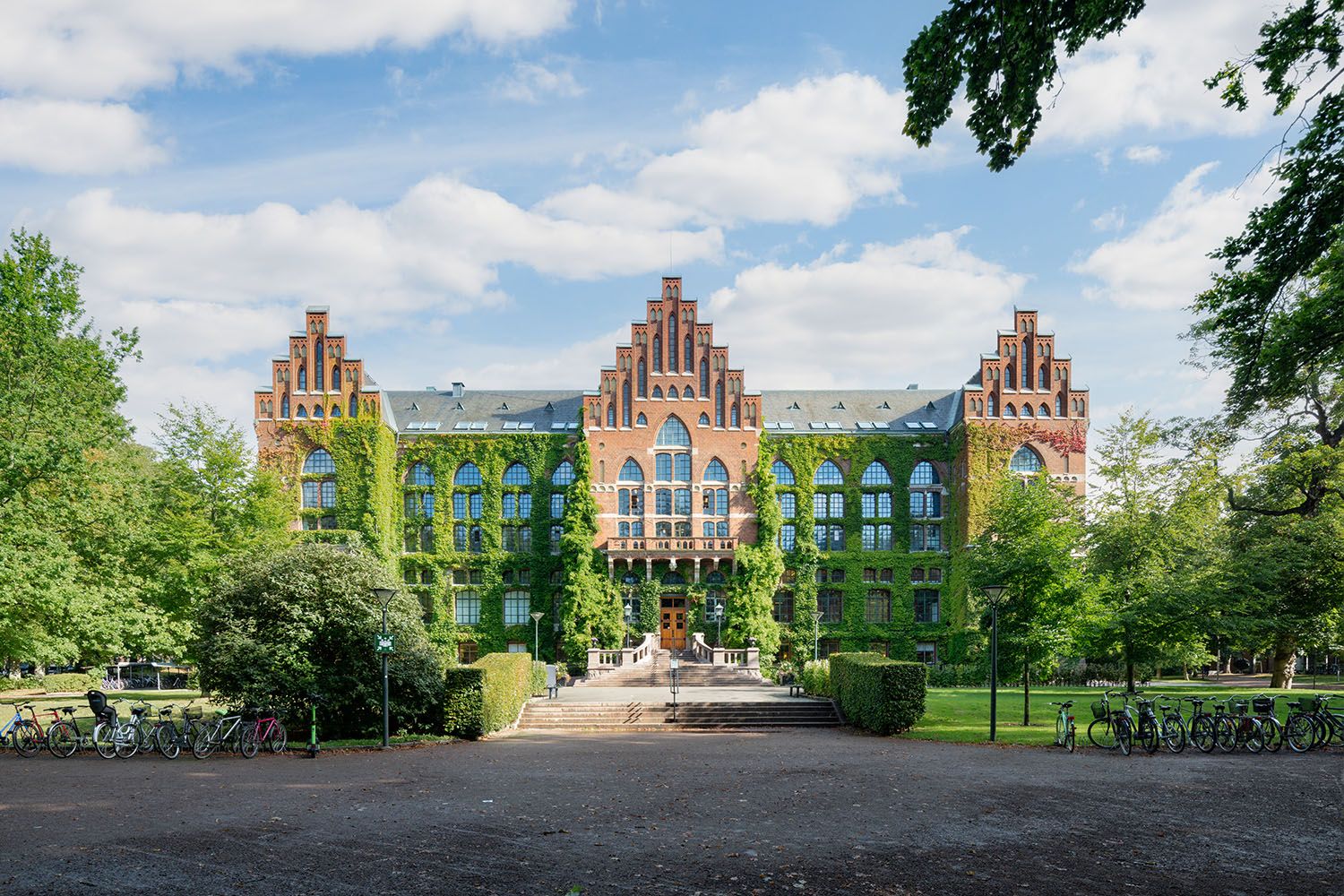 A large, ivy-covered brick building with gabled roofs, in a green park with trees and parked bicycles.