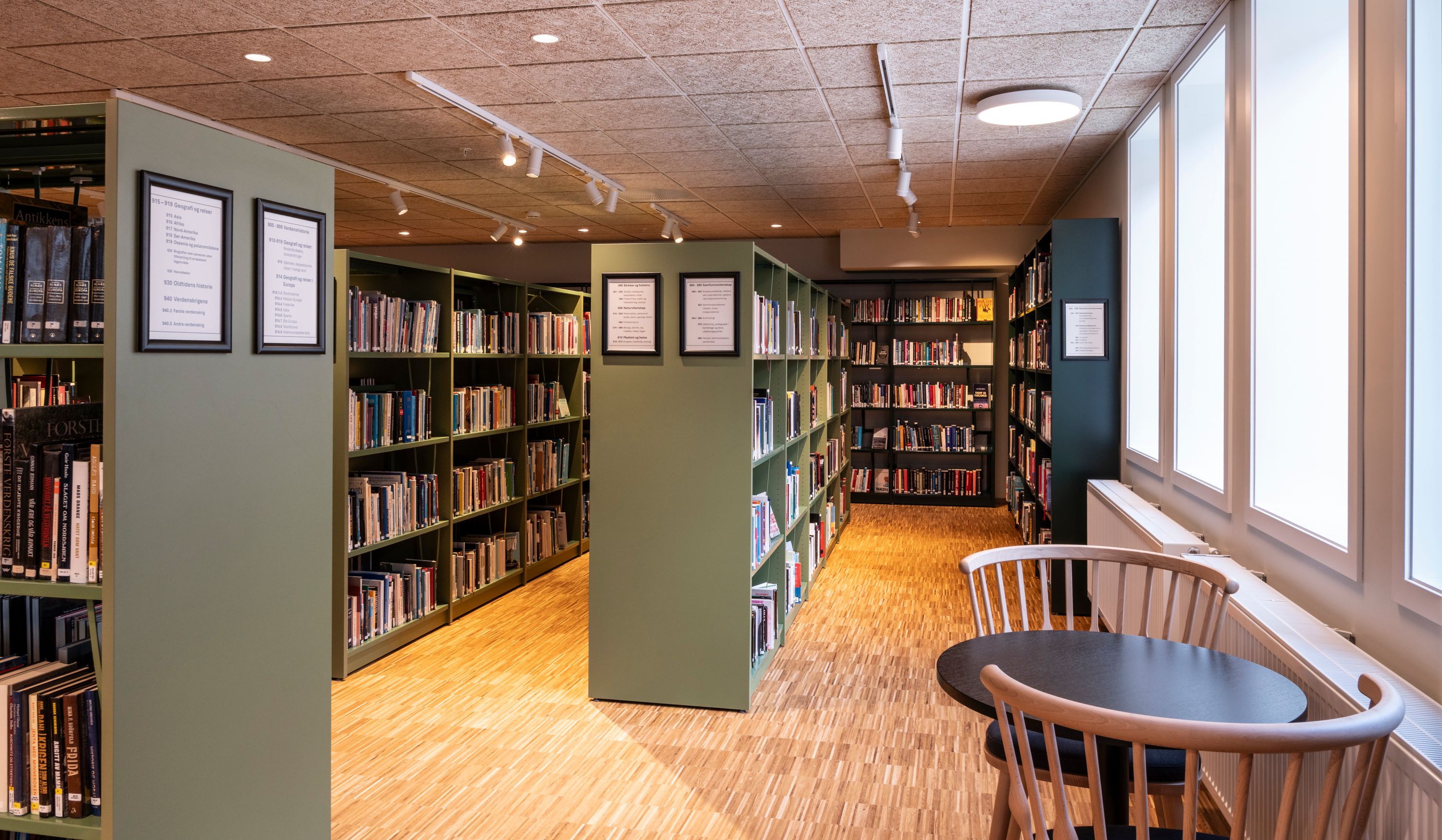 A modern library with green bookshelves, a wooden floor, and a small table with two chairs by tall windows.