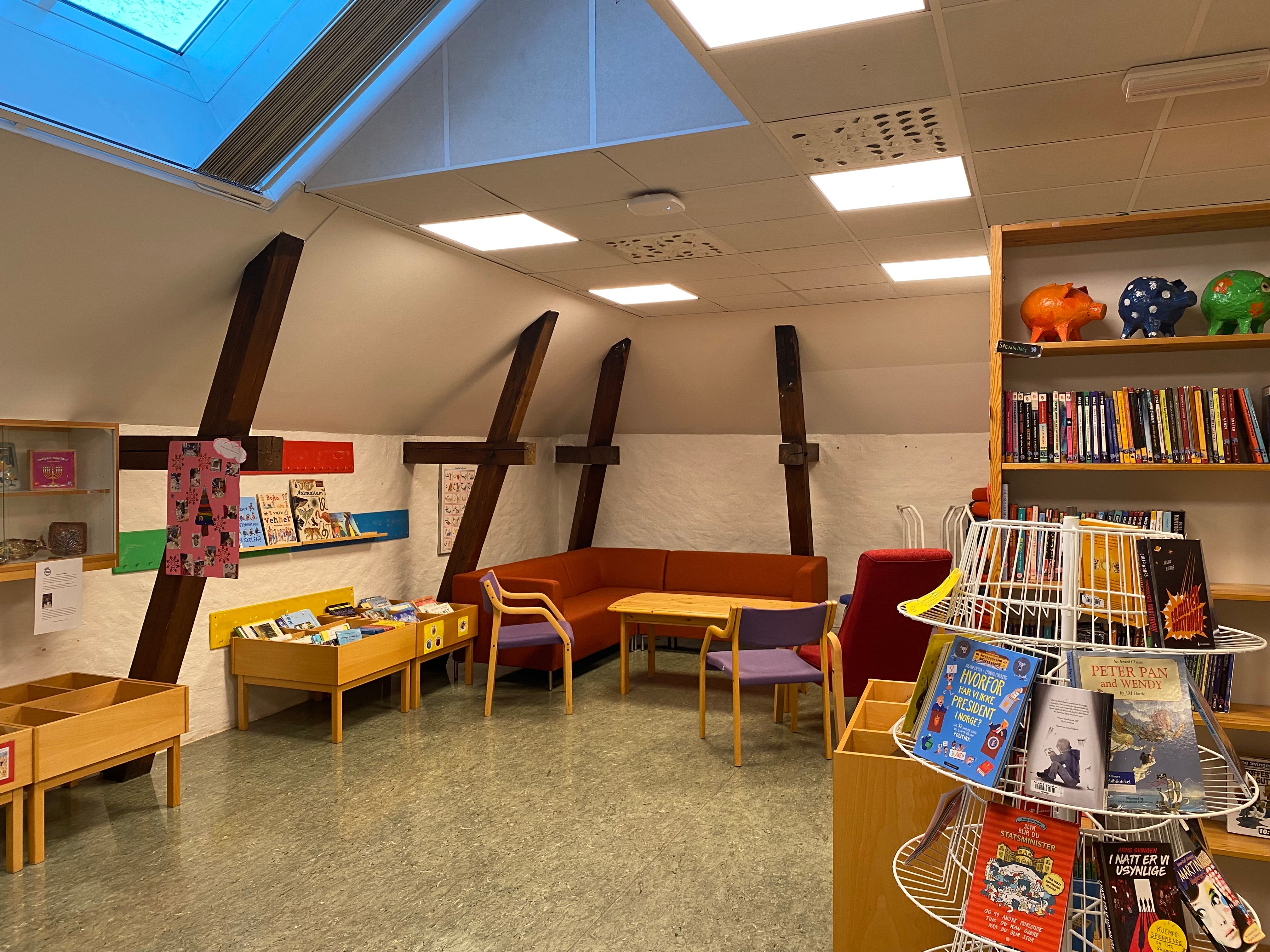 An attic children's library with a skylight, exposed wooden beams, an orange couch, and numerous bookshelves filled with books.