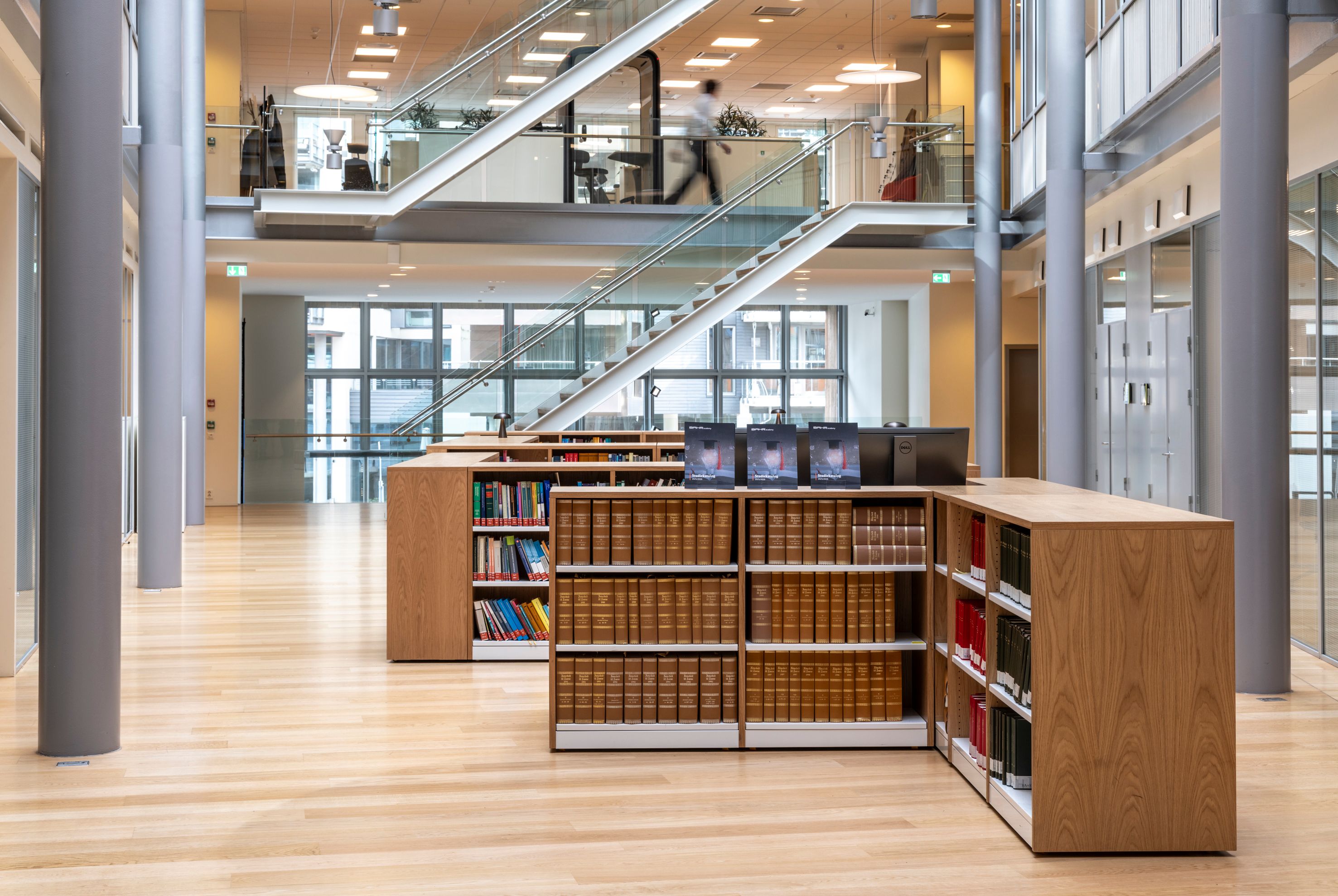 Modern, spacious library with light wooden floors, low bookshelves, a prominent glass staircase, and a person on the upper level.