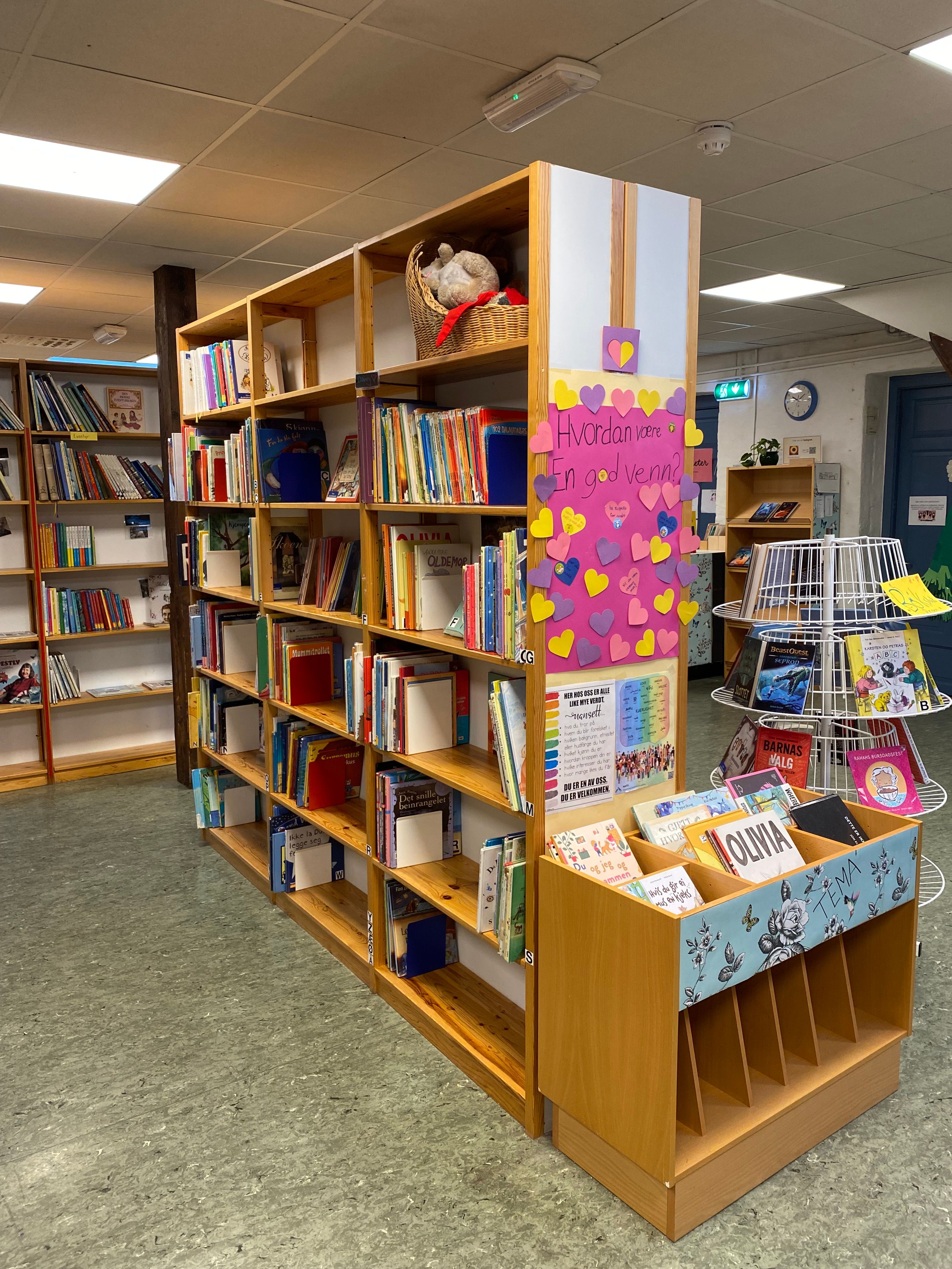 A children's library with wooden bookshelves full of books, a pink display decorated with colorful hearts, and various book stands.