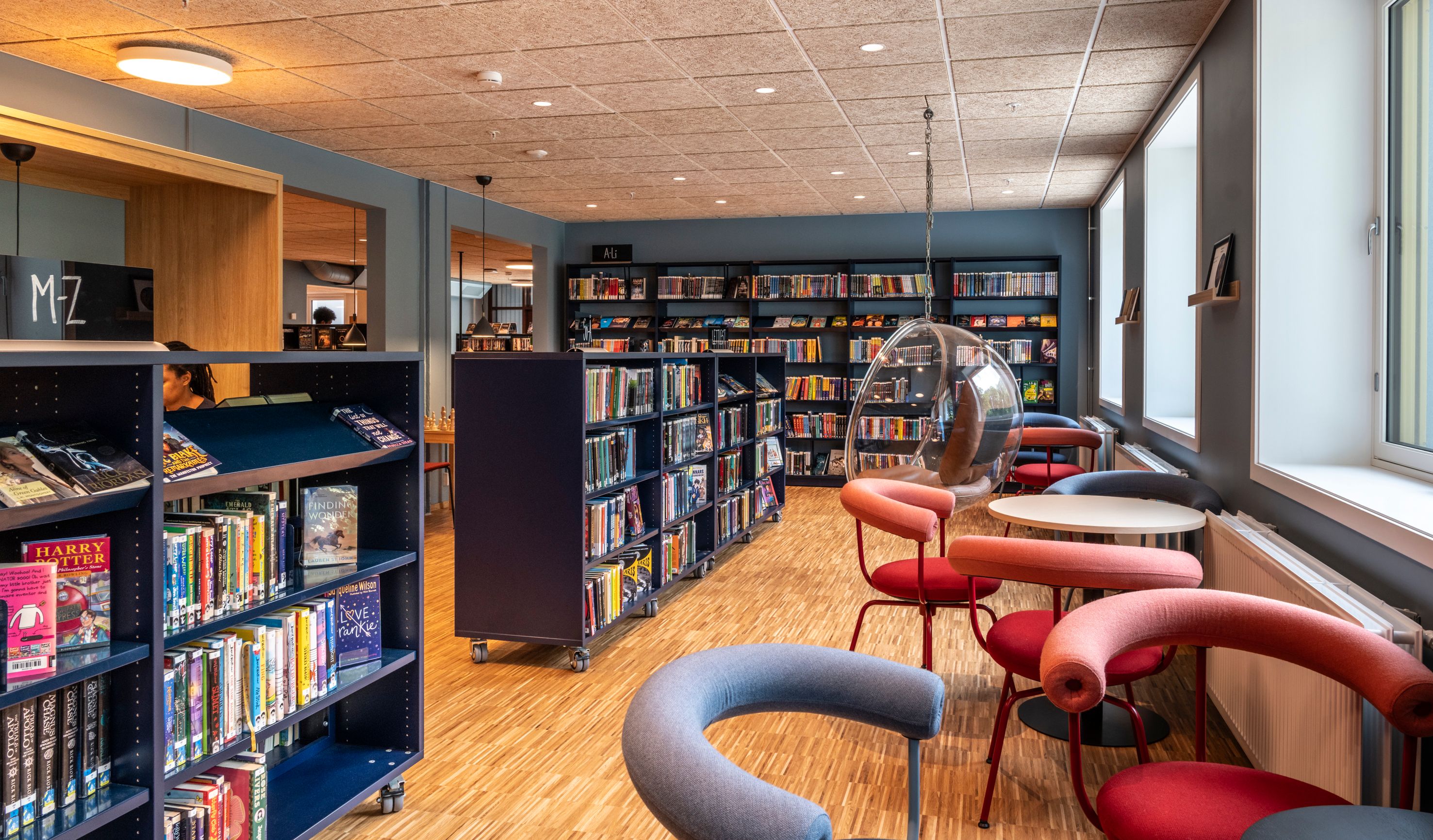 A modern library with bookshelves, colorful chairs, a clear hanging bubble chair, and windows along the right wall.