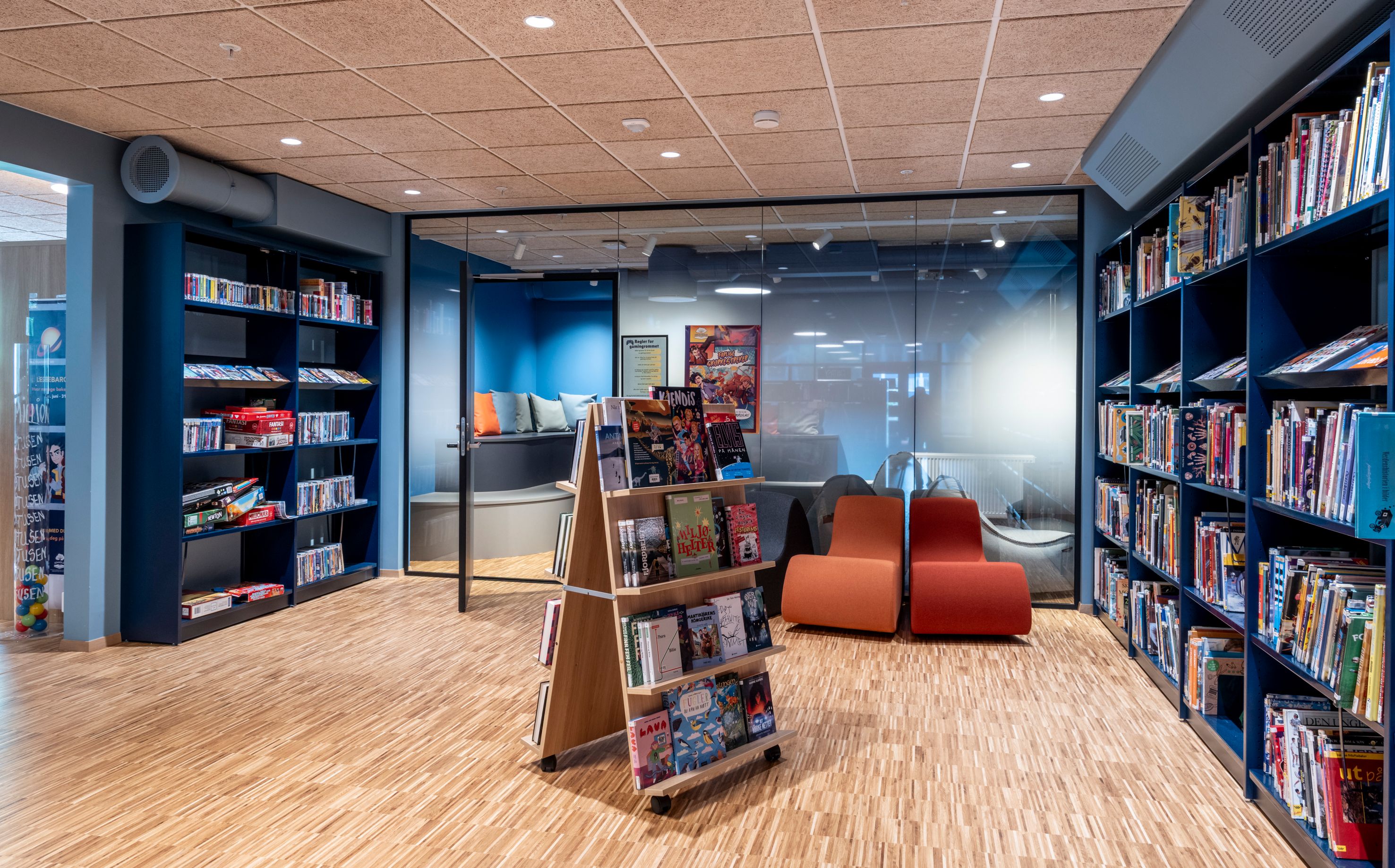 Modern library interior with blue bookshelves, a wooden floor, orange lounge chairs, and a glass-walled quiet room.