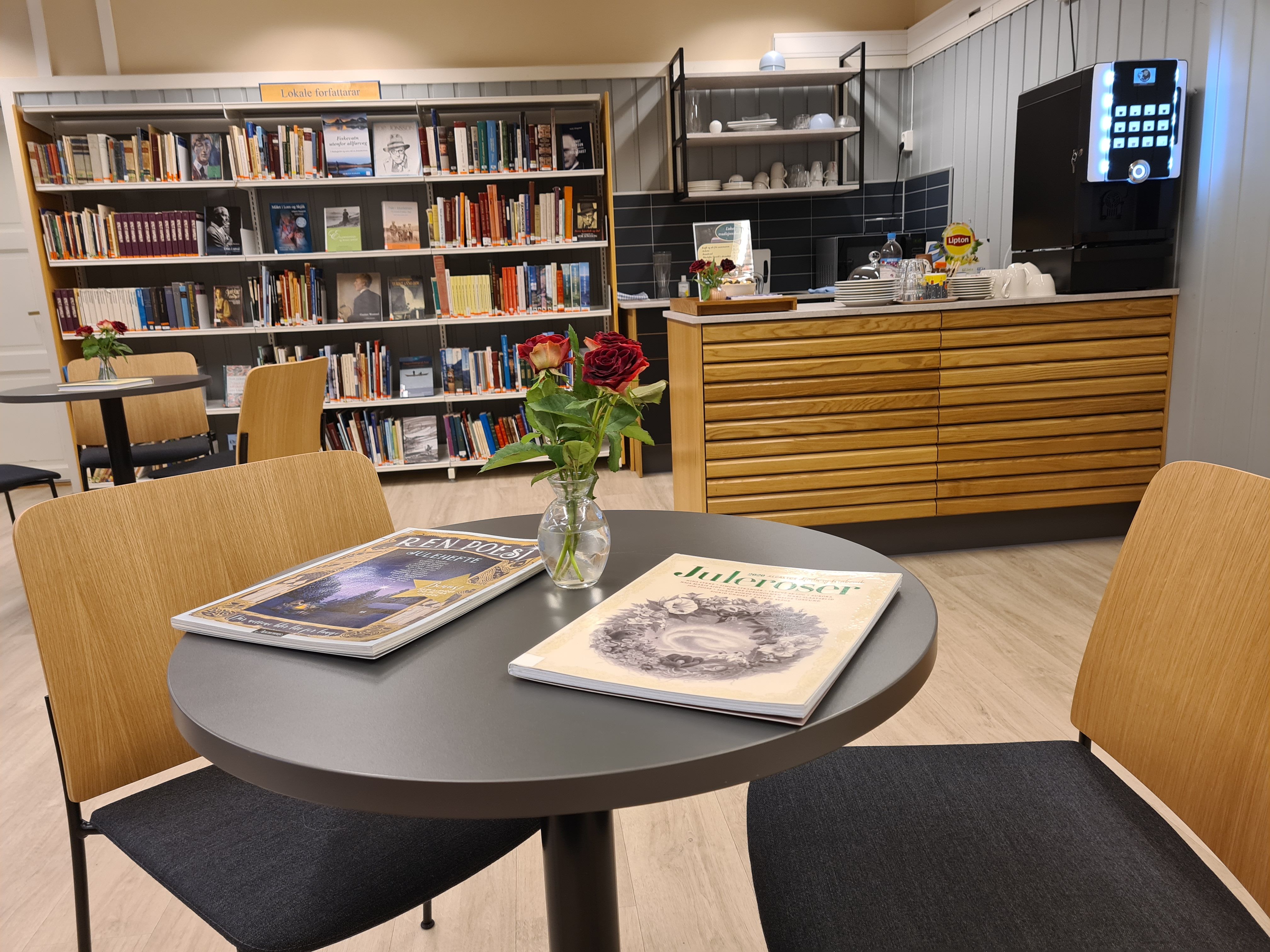 A table with magazines and red roses sits in a library lounge area with bookshelves and a coffee station.