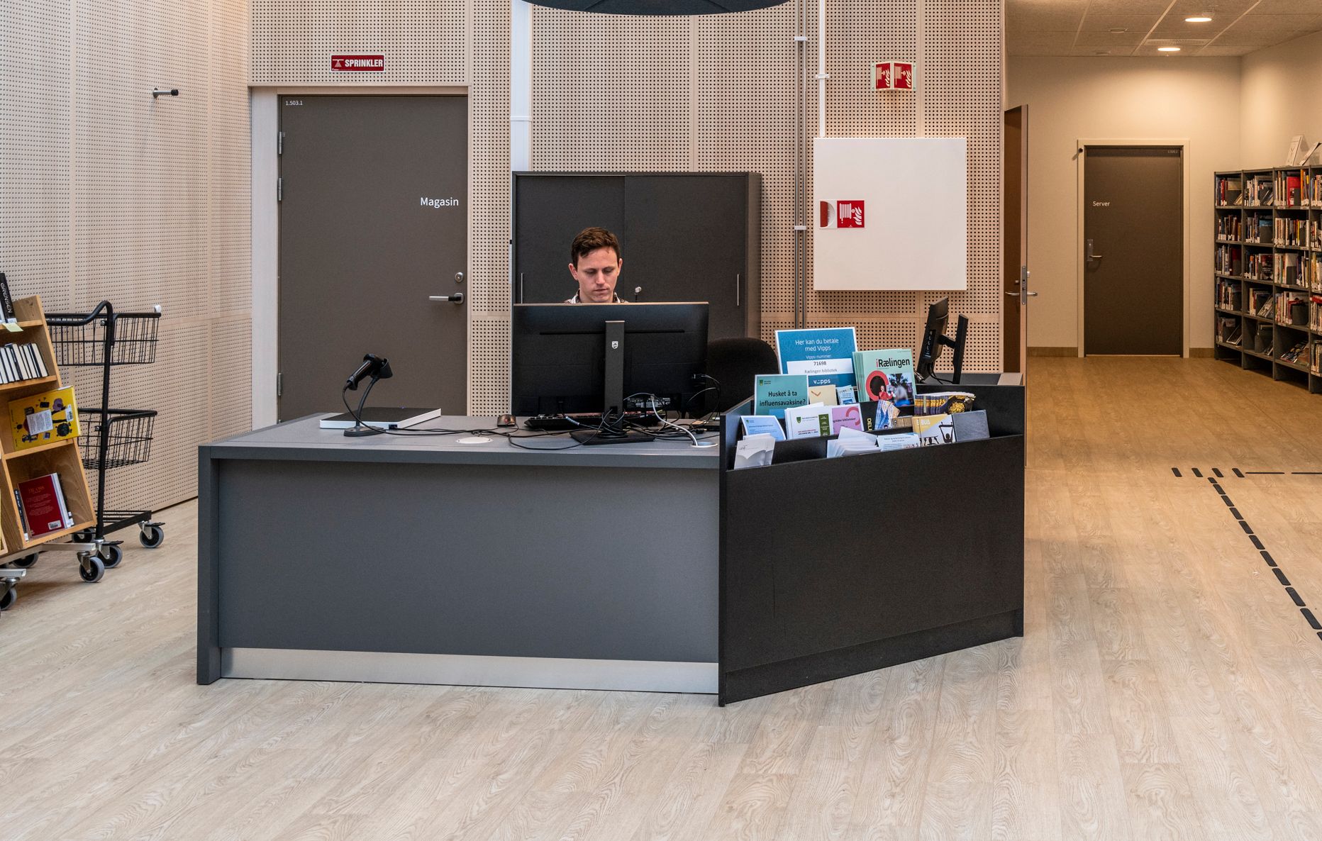 en mann sitter ved en grå bibliotekskranke og jobber på en pc