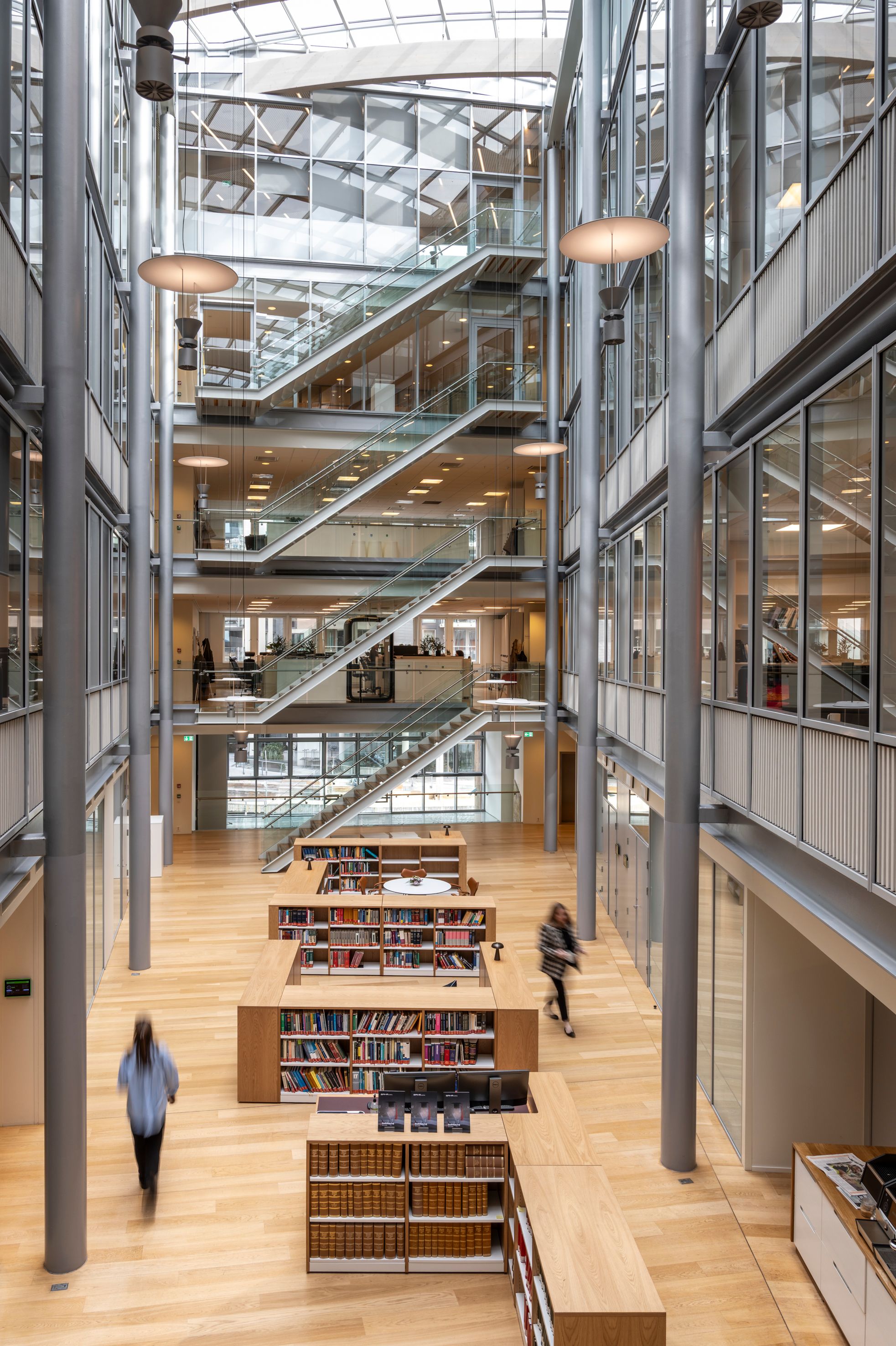 Modern multi-story library atrium with wooden floors, glass staircases, bookshelves, and offices visible through glass walls.