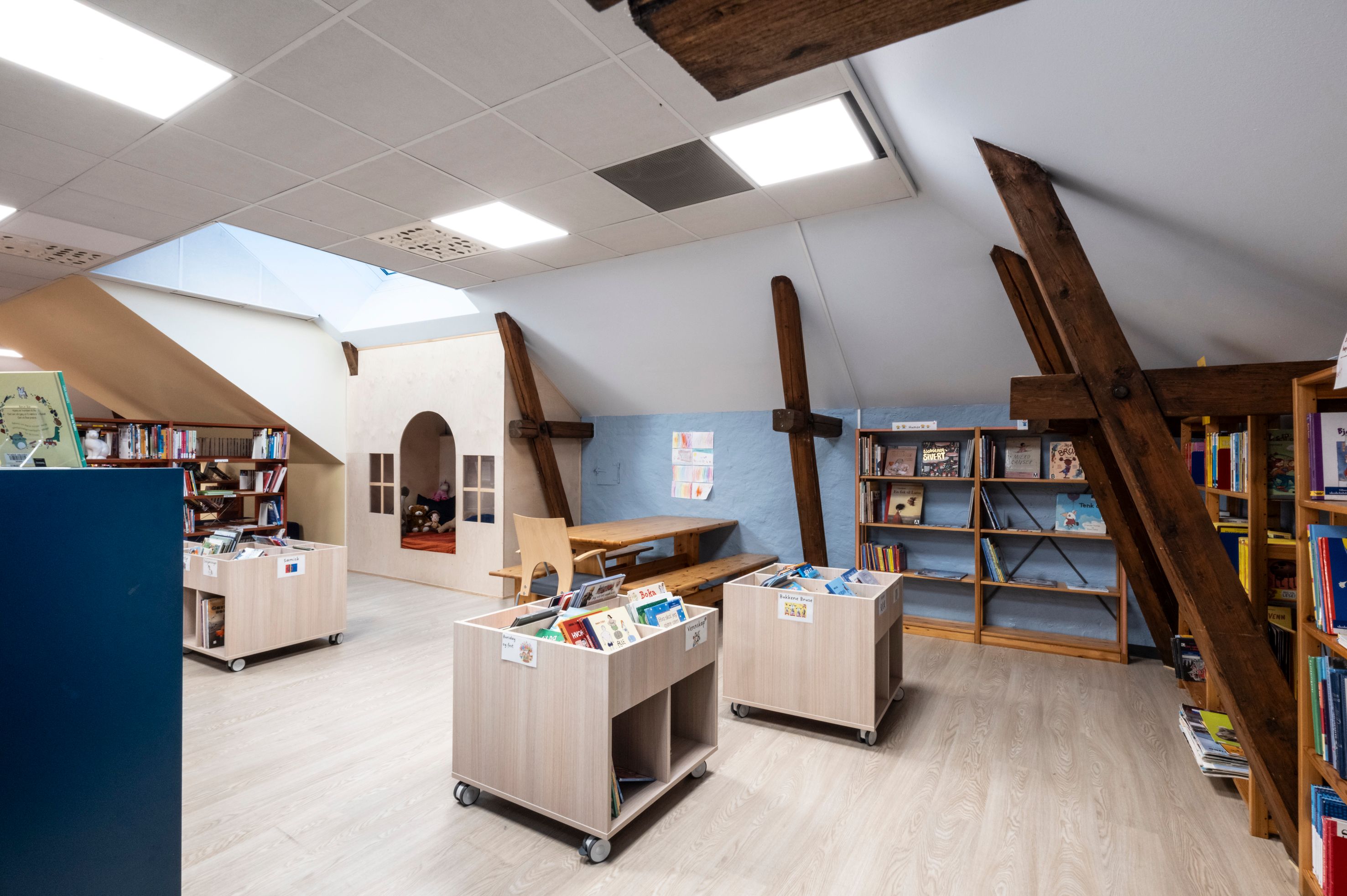 A bright, modern attic library with bookshelves, a cozy reading nook, and exposed wooden beams.