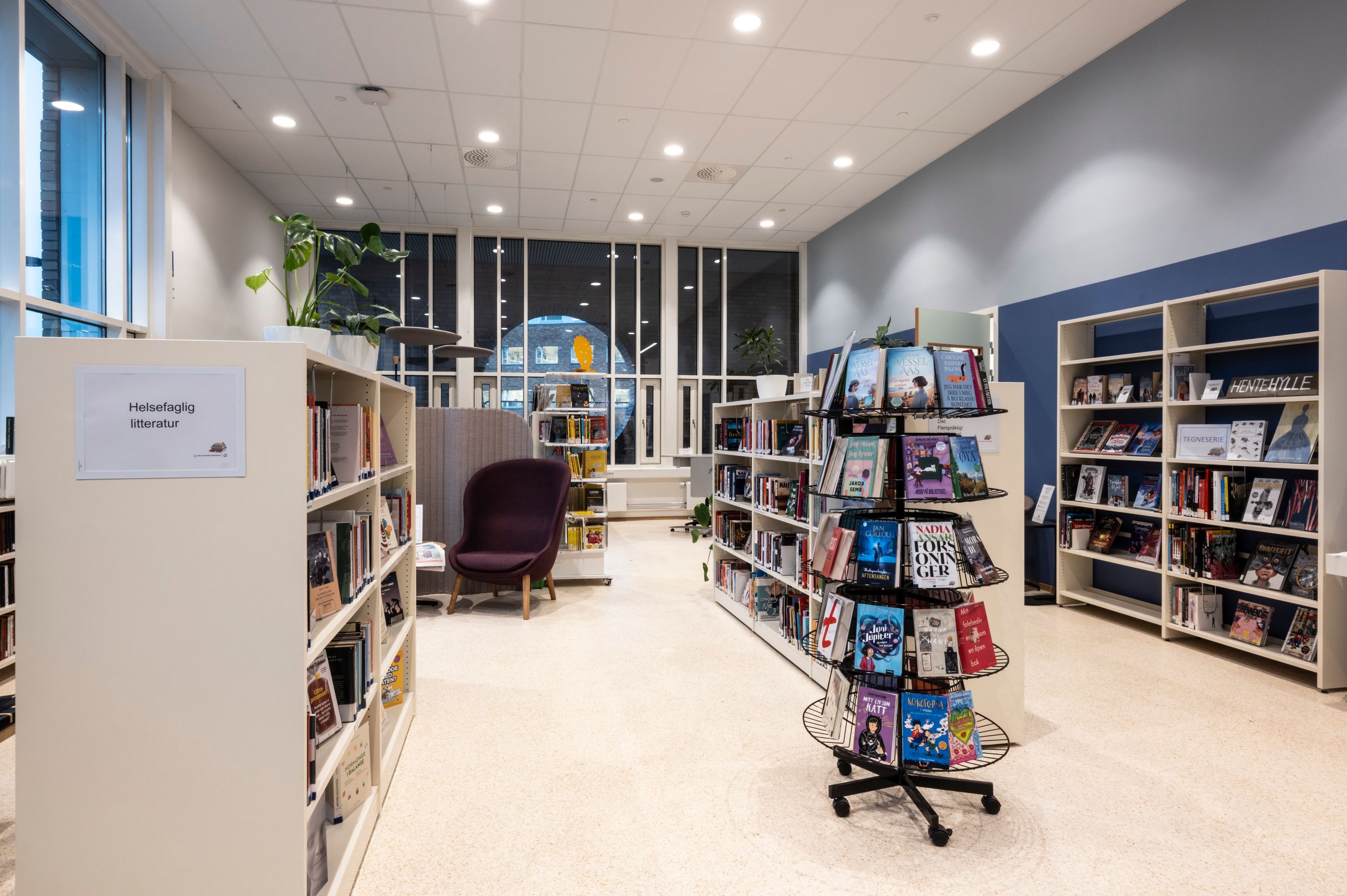 A brightly lit modern library with rows of bookshelves, a rotating book display, and a purple armchair by large windows.