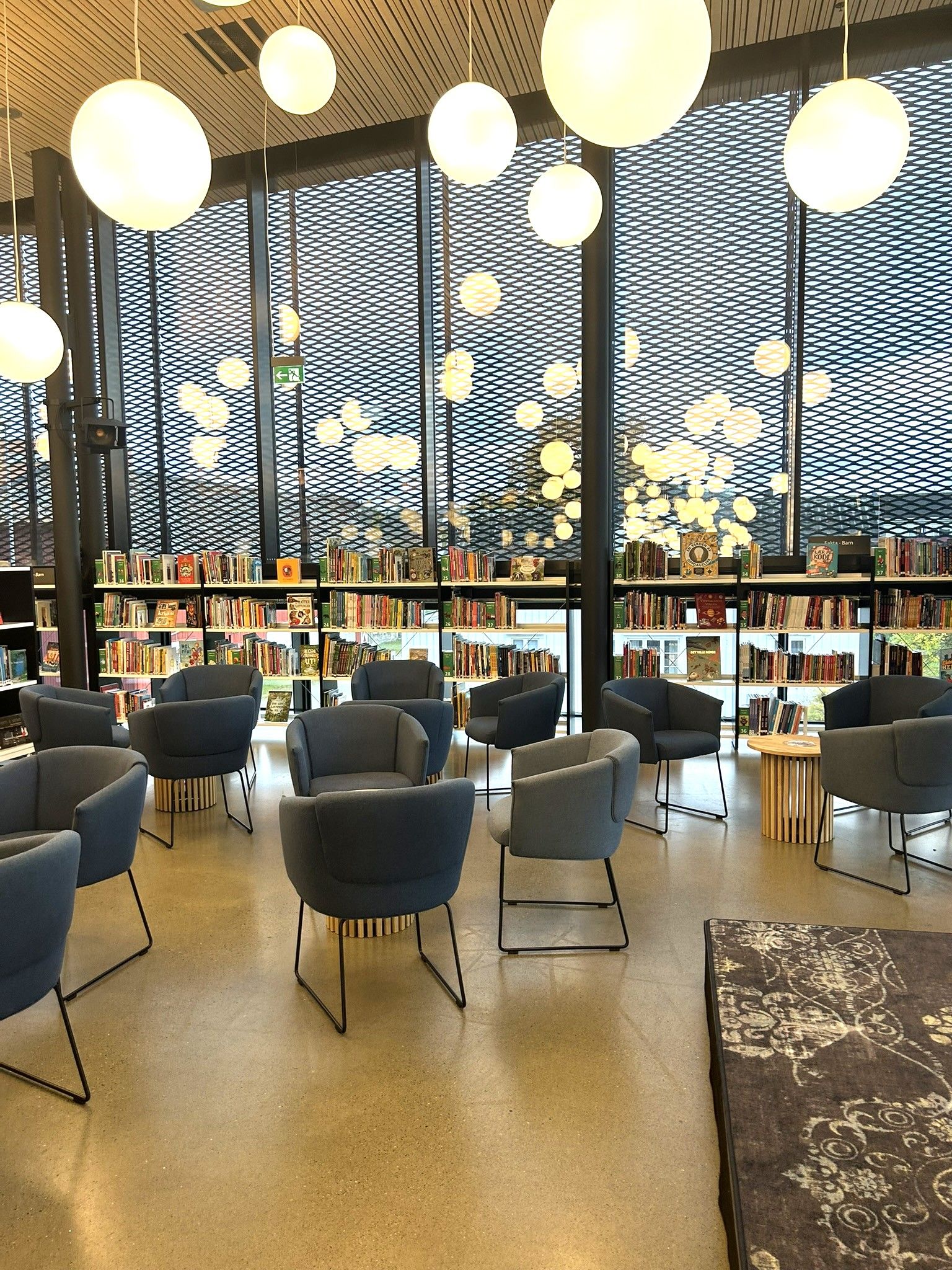 A modern library reading area with blue armchairs, bookshelves, and large windows featuring a dark mesh and glowing spherical lights.