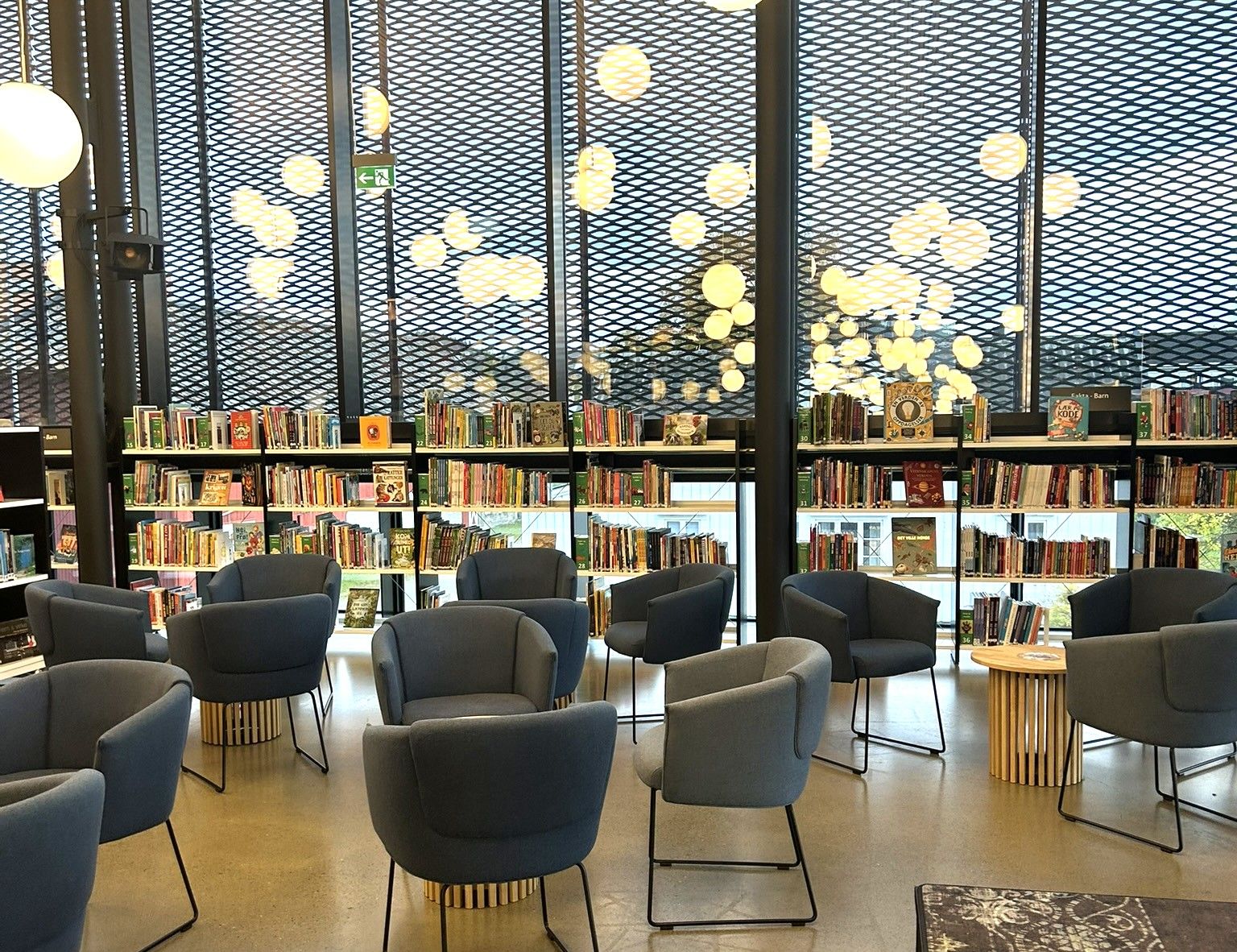 A modern library reading area with blue armchairs, bookshelves, and large windows featuring a dark mesh and glowing spherical lights.