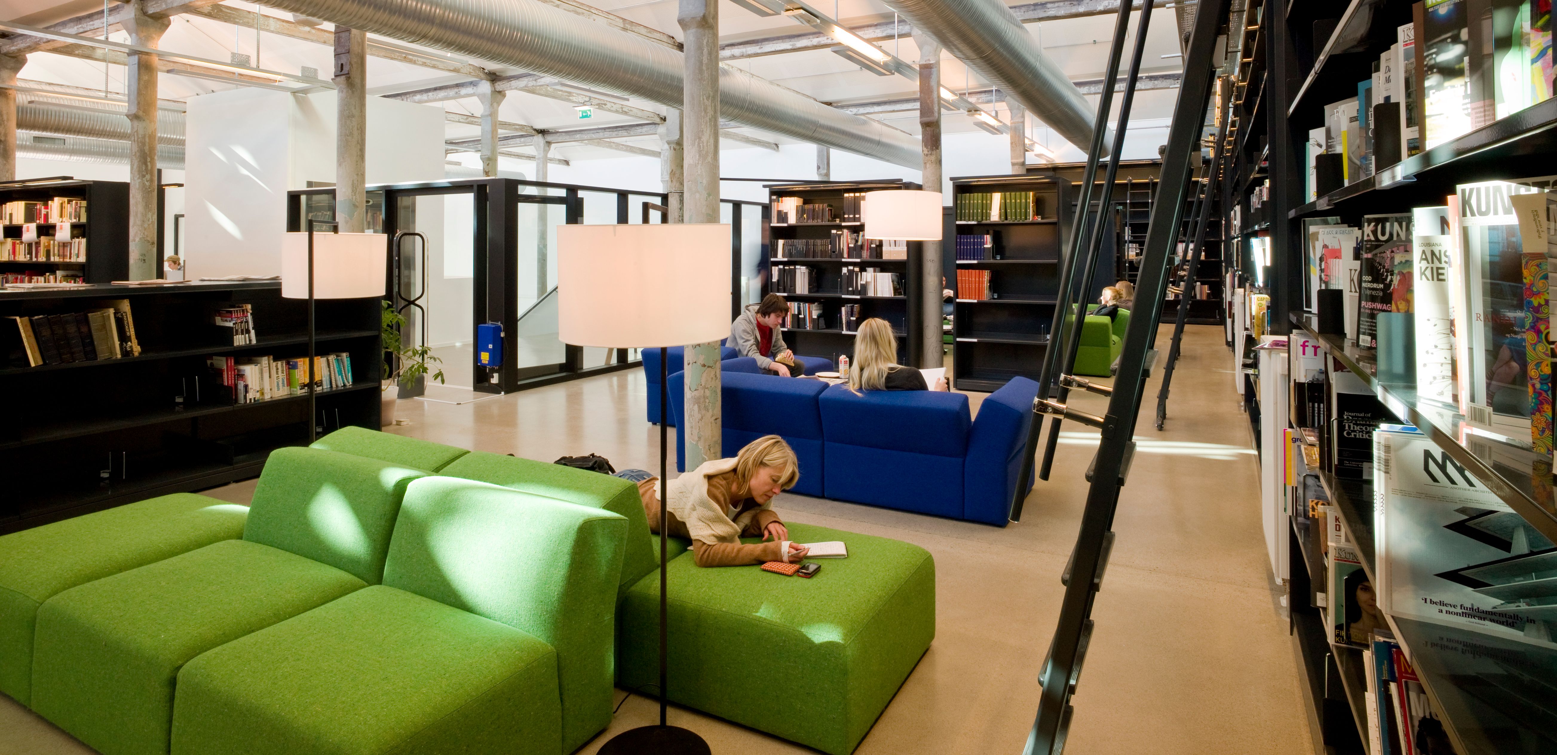 a woman is reading a book in a library while sitting on a green couch .