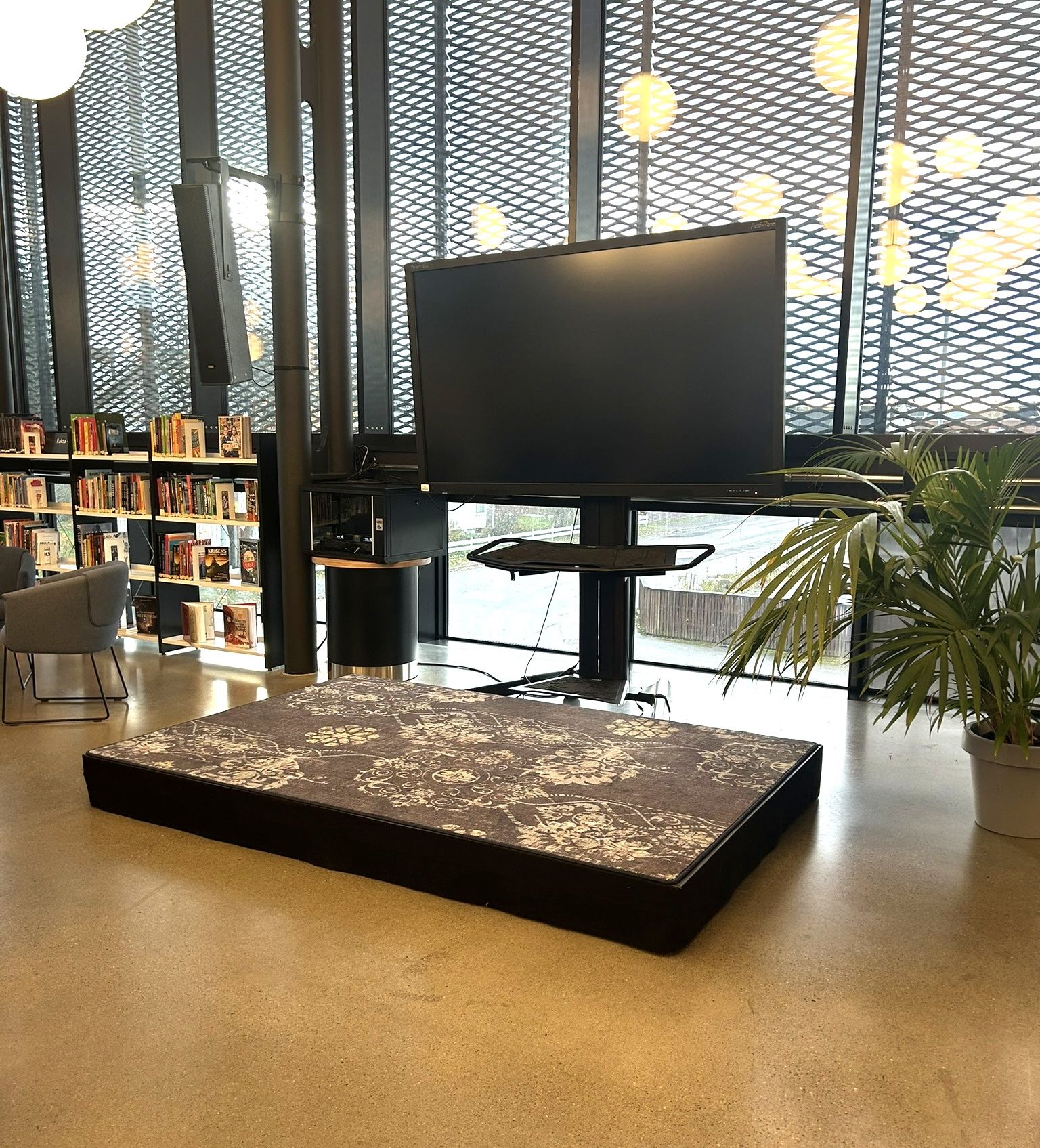 A modern library or study room with a large TV screen, a low rectangular patterned mat, bookshelves, and large windows.