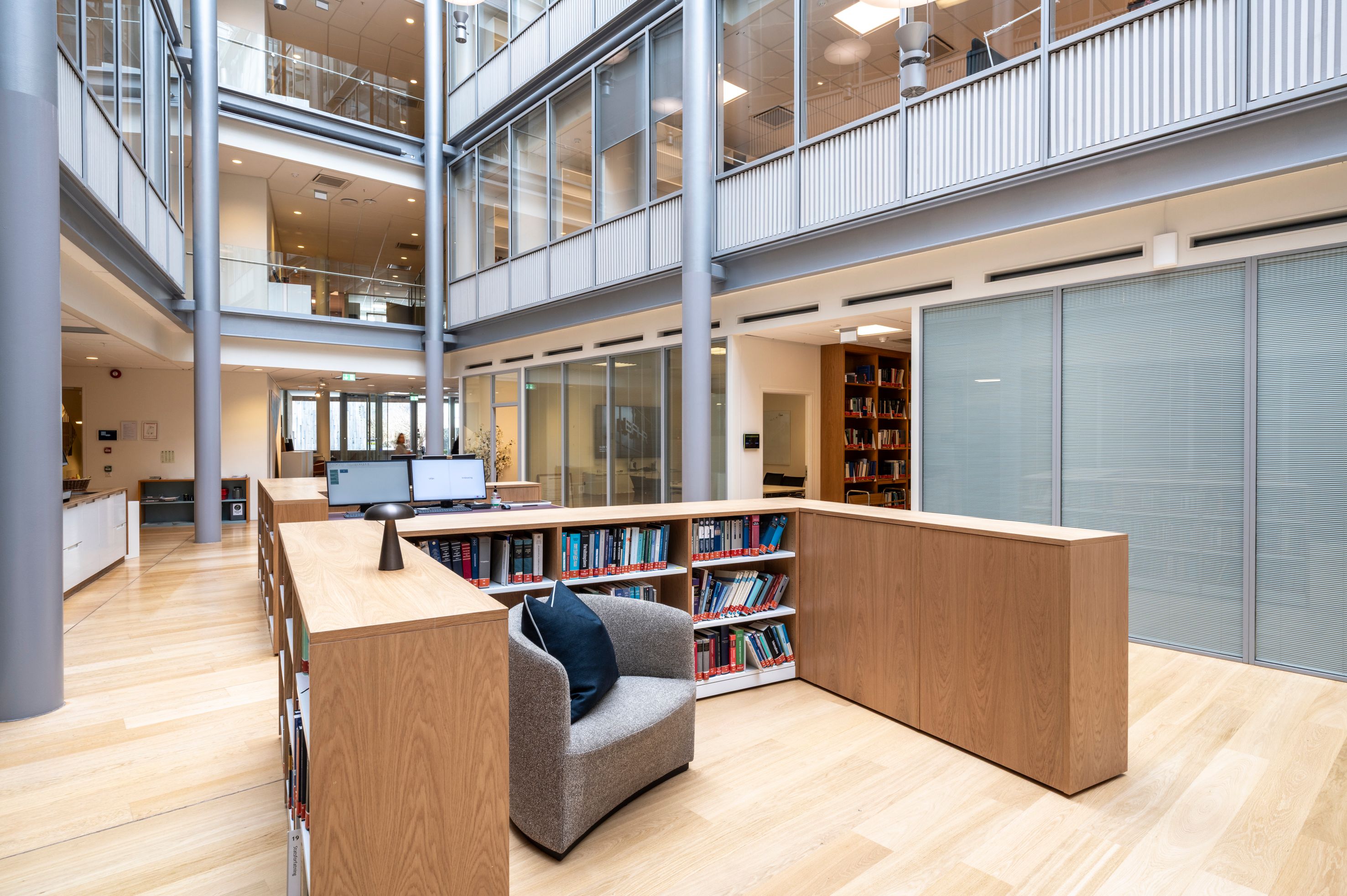 A modern library and study area with light wood shelves and seating, in a multi-story glass atrium.