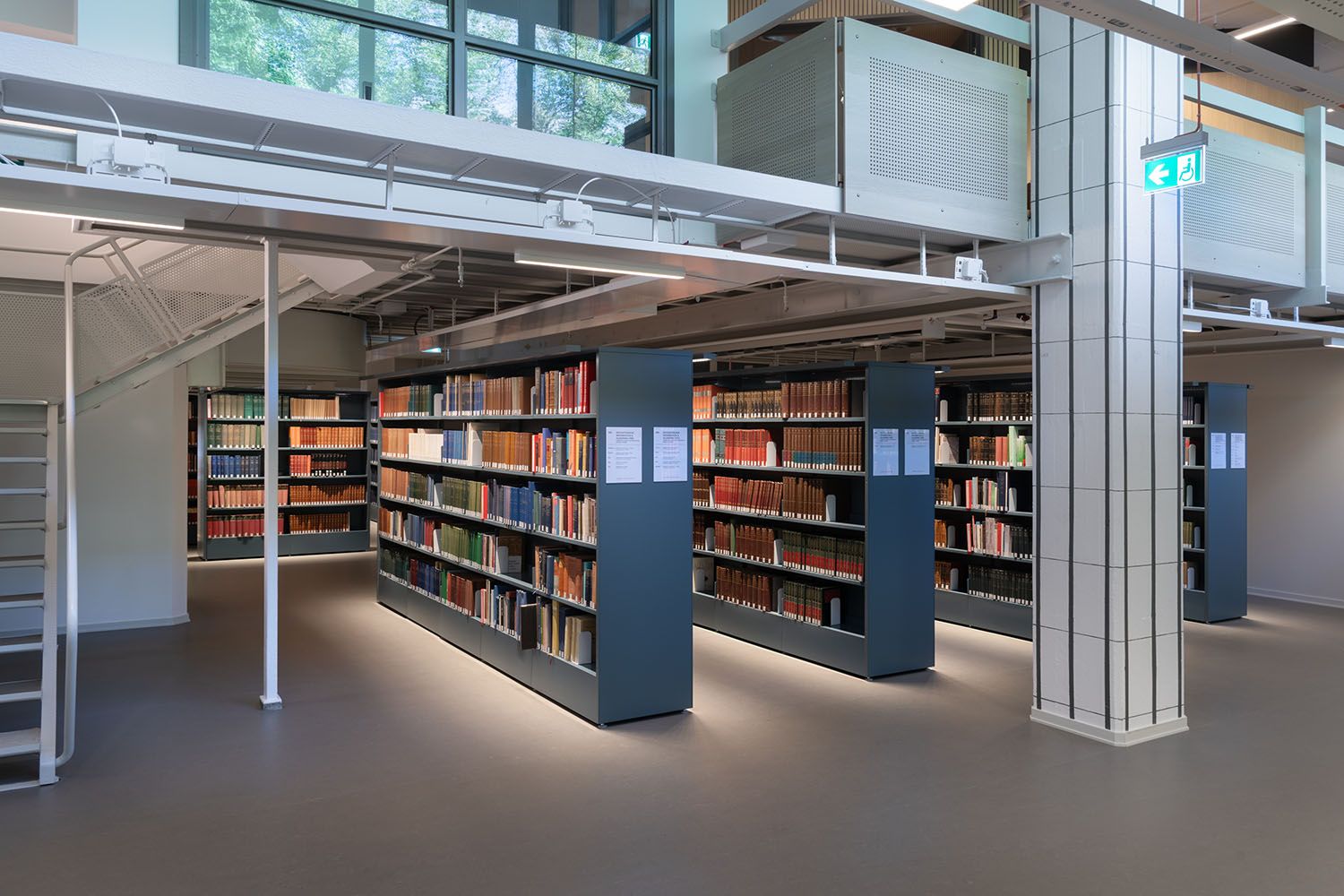A modern library interior with rows of dark bookshelves filled with books, bright lighting, a staircase, and large windows.