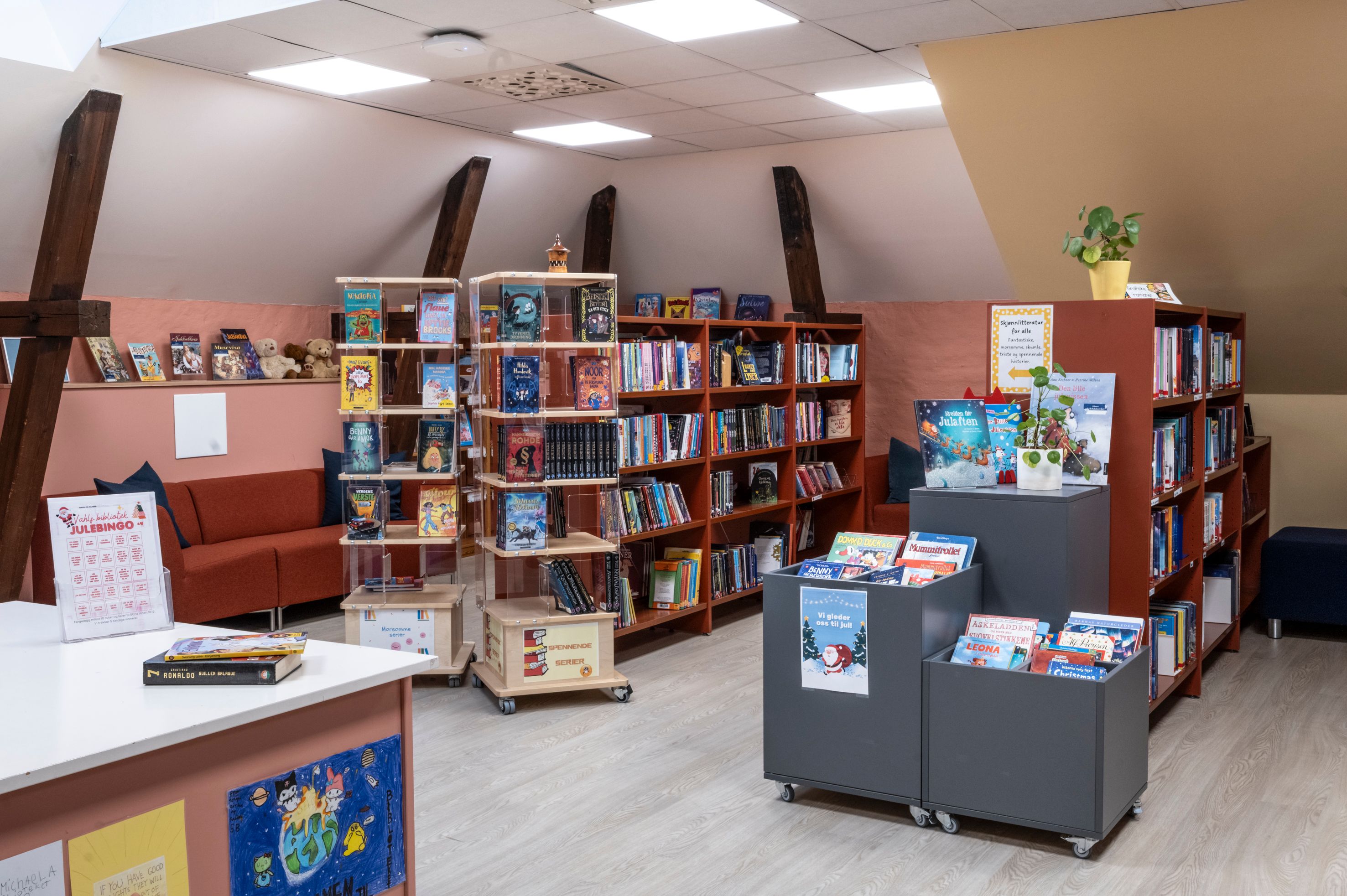 An inviting children's library with numerous books on shelves and display units, a red sofa, and exposed wooden ceiling beams.