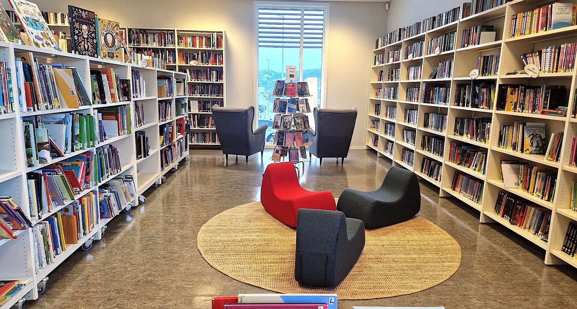 Modern library with white bookshelves filled with books, a seating area with red and dark gray lounge chairs on a round rug, and two armchairs.