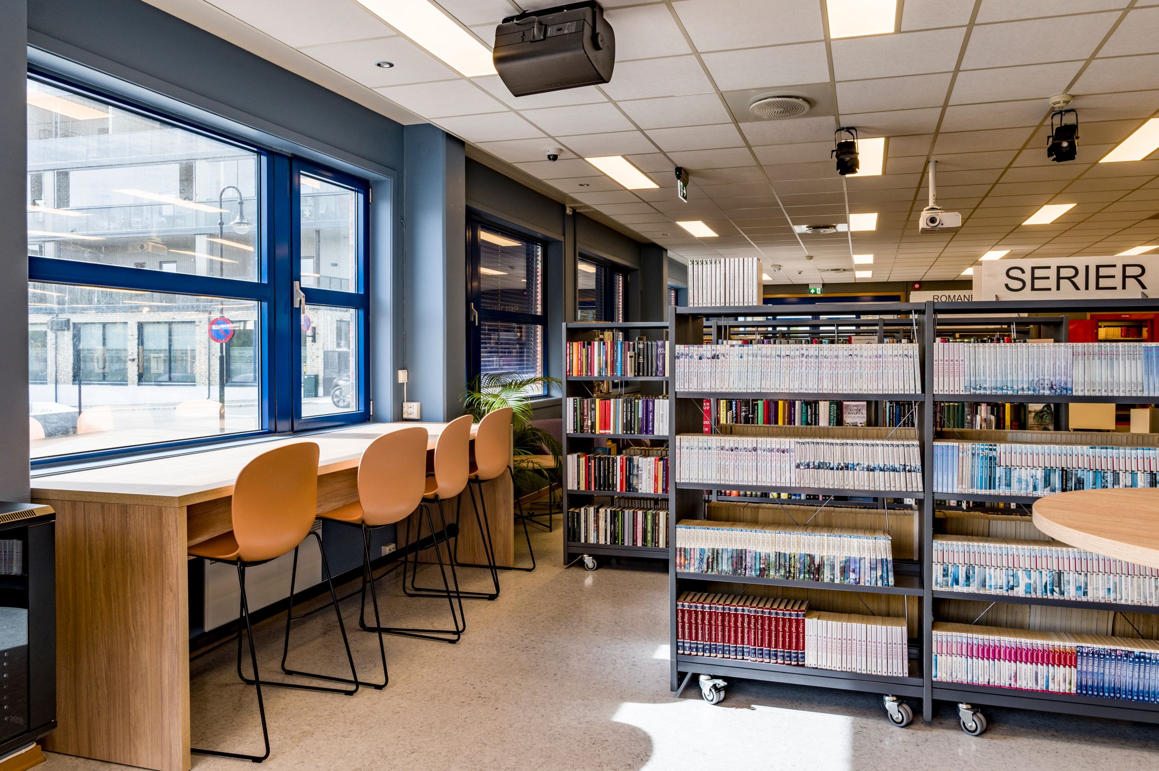 et bibliotek med mange bokreoler og en lang bordrekke med høye stoler foran vinduene