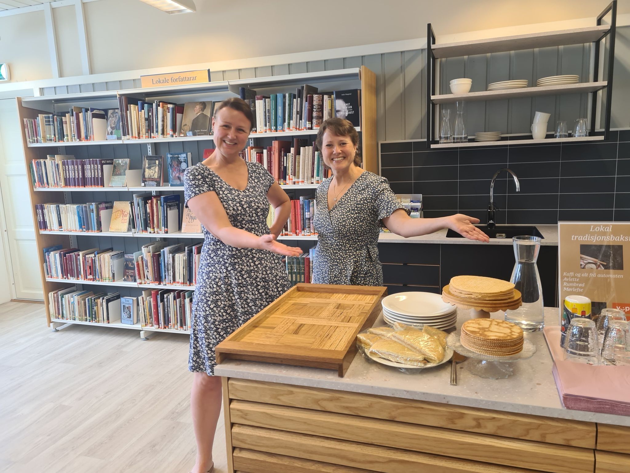 Two smiling women gesture towards a counter laden with traditional baked goods and plates, set against a backdrop of library shelves and a kitchen.