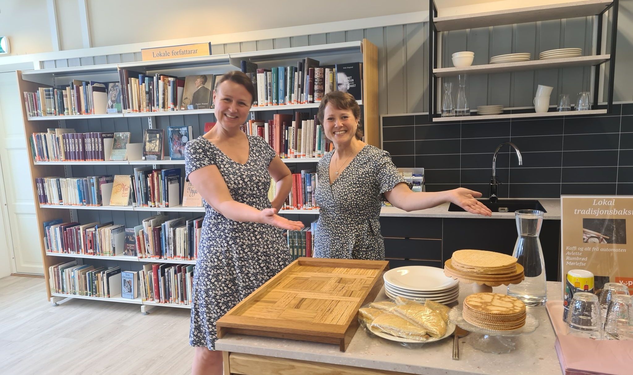 Two smiling women stand in a library cafe, gesturing towards a counter displaying traditional Norwegian pancakes.