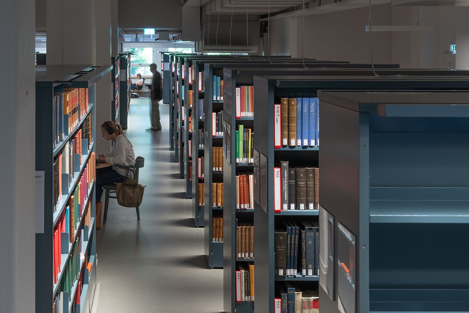 A person studies at a desk in a library lined with rows of bookshelves.