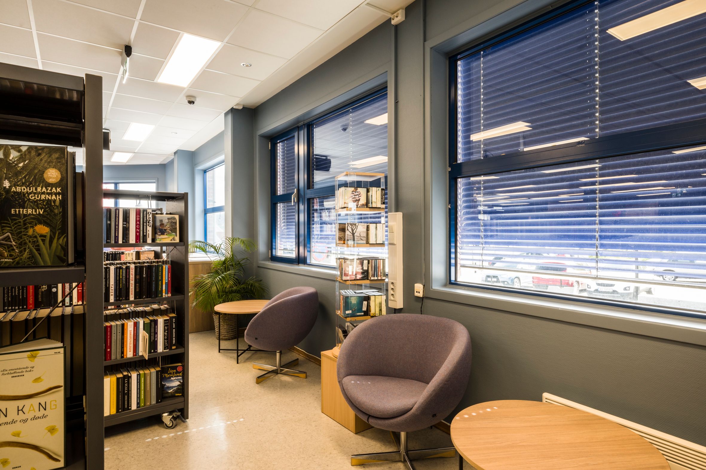 et bibliotek med bokreoler på hjul og 2 lave stoler og bord foran vinduene