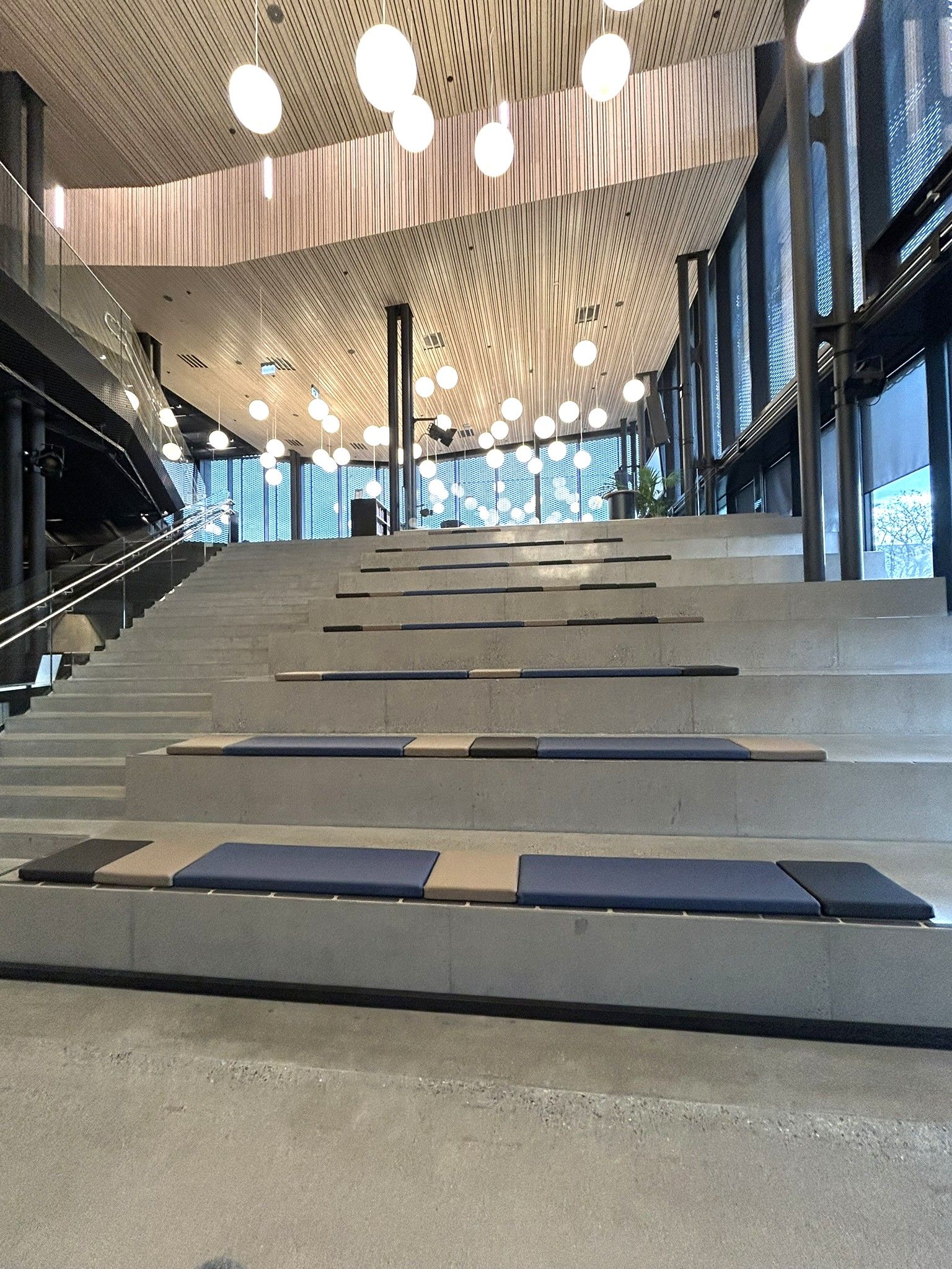 Modern tiered concrete seating and stairs with blue and tan cushions under a wood-paneled ceiling with numerous spherical pendant lights.