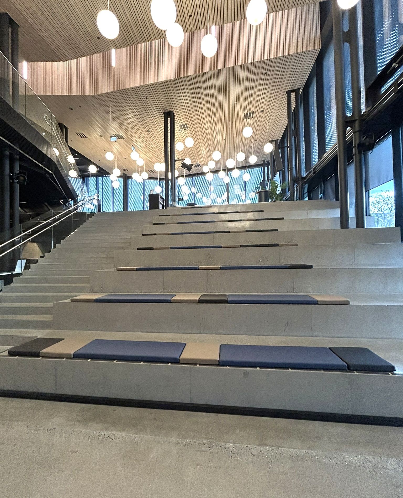 Modern tiered concrete seating and stairs with blue and tan cushions under a wood-paneled ceiling with numerous spherical pendant lights.