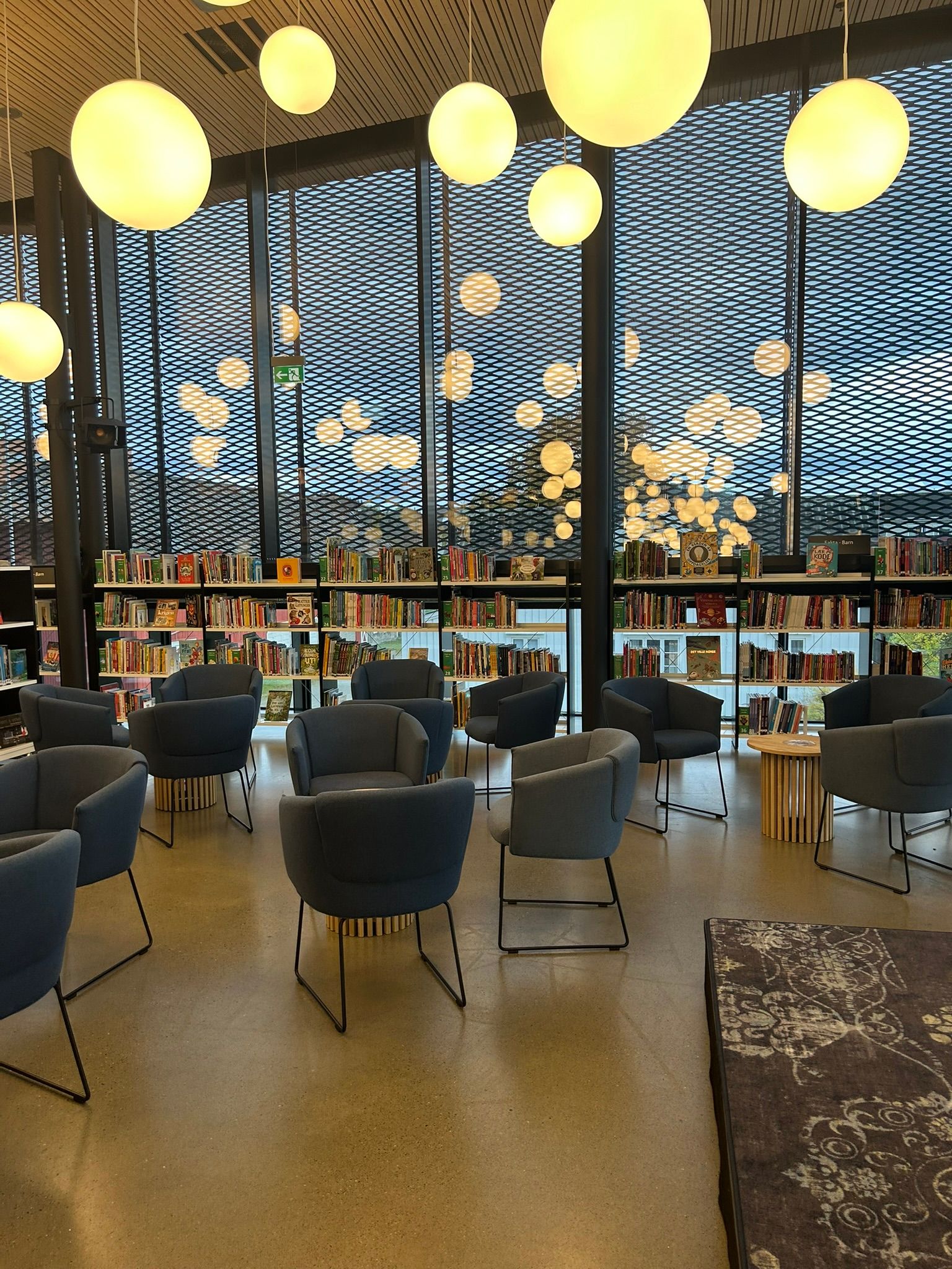 A modern library with grey armchairs, bookshelves, and large windows with circular lights.