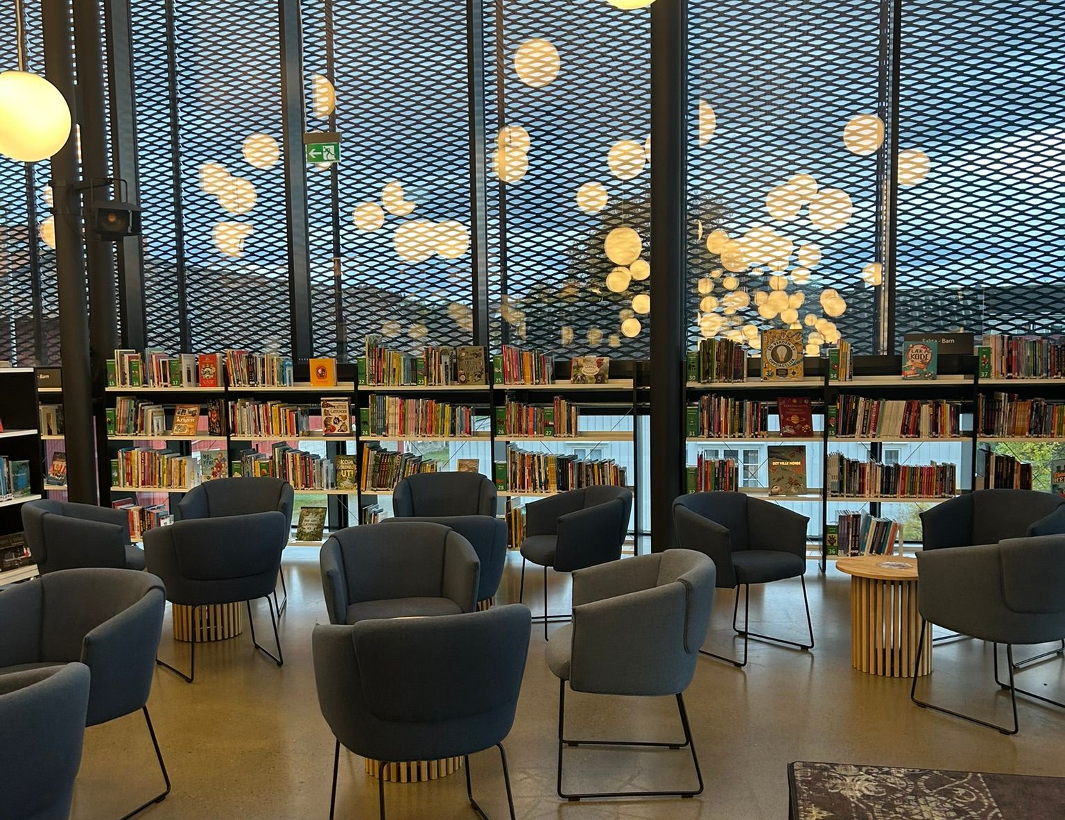 A modern library with grey armchairs, bookshelves, and large windows with circular lights.