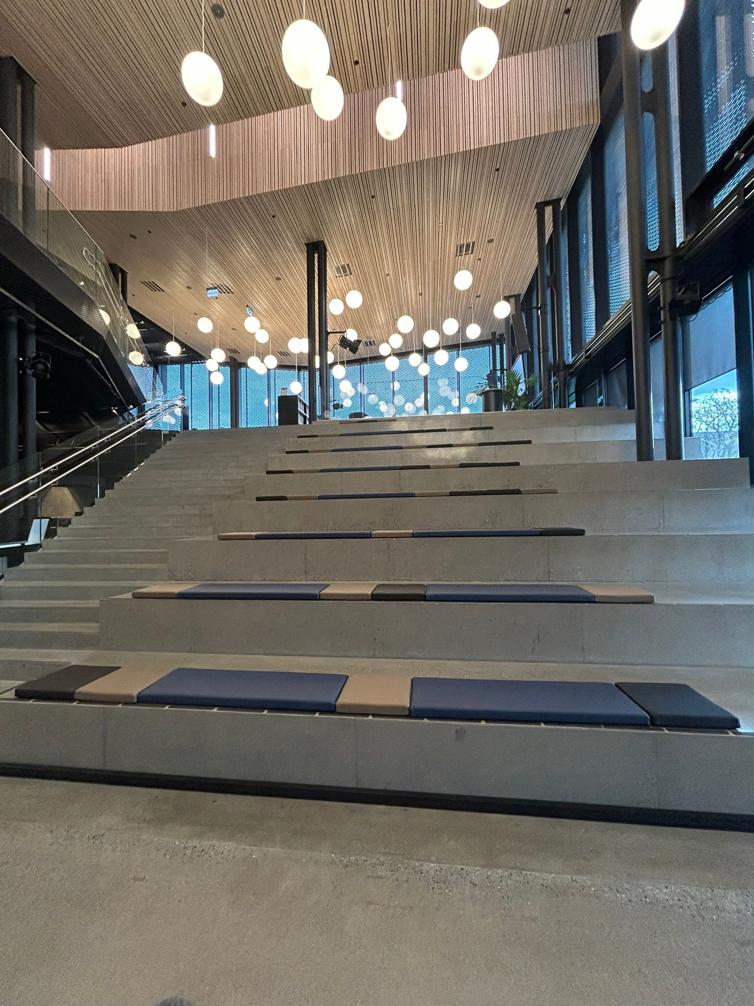 Modern hall with wide concrete stairs for seating, a wooden ceiling, and spherical pendant lights.