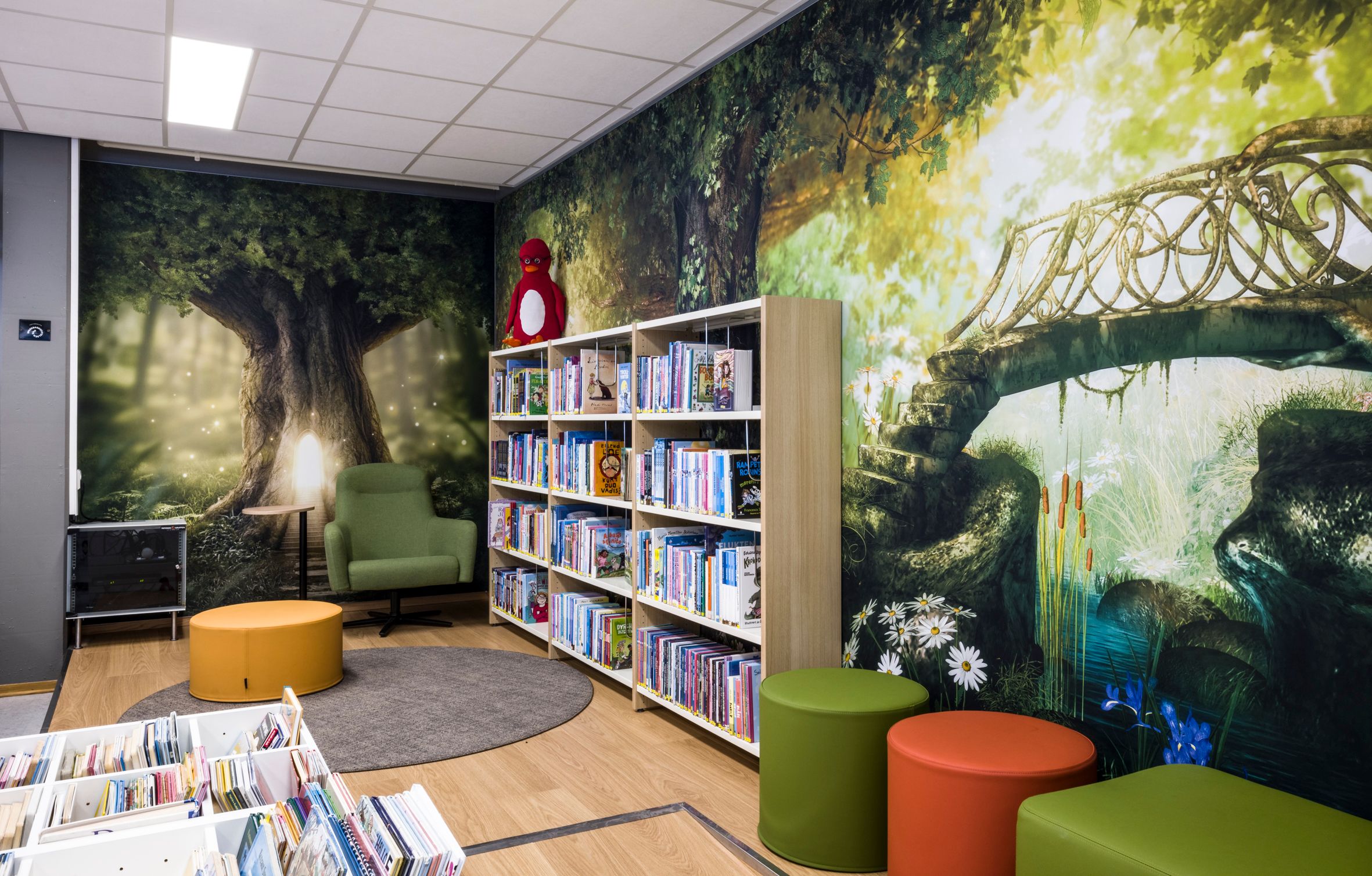 A whimsical children's library with forest mural walls, bookshelves, and colorful seating.