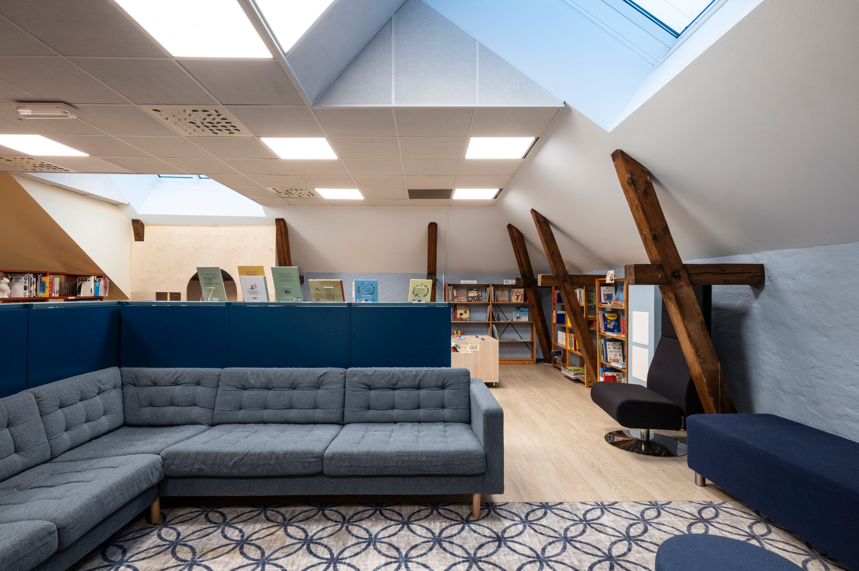 Modern attic library with a blue sectional sofa, skylights, and exposed wooden beams.