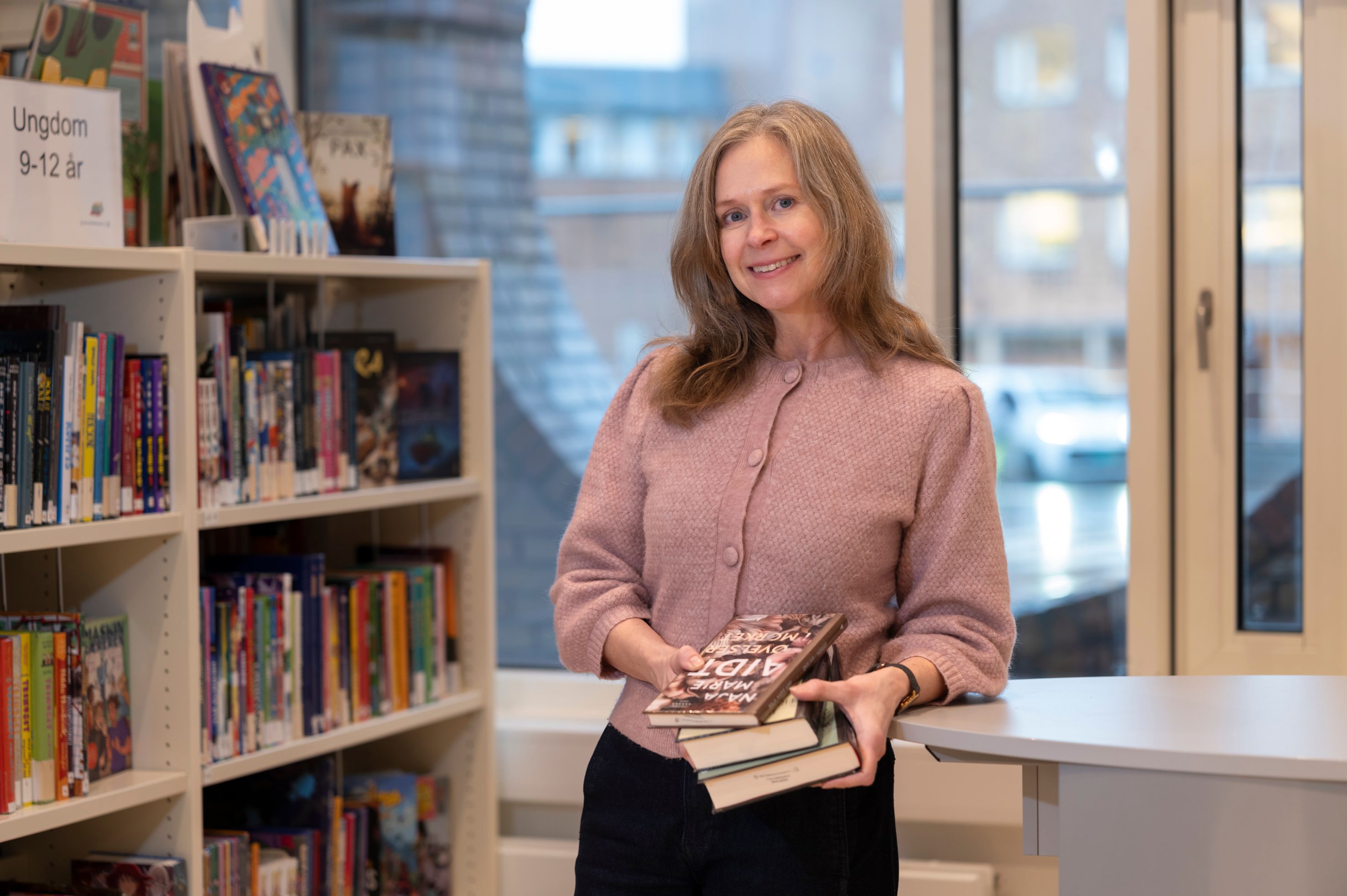 A smiling woman holds a stack of books in a library.