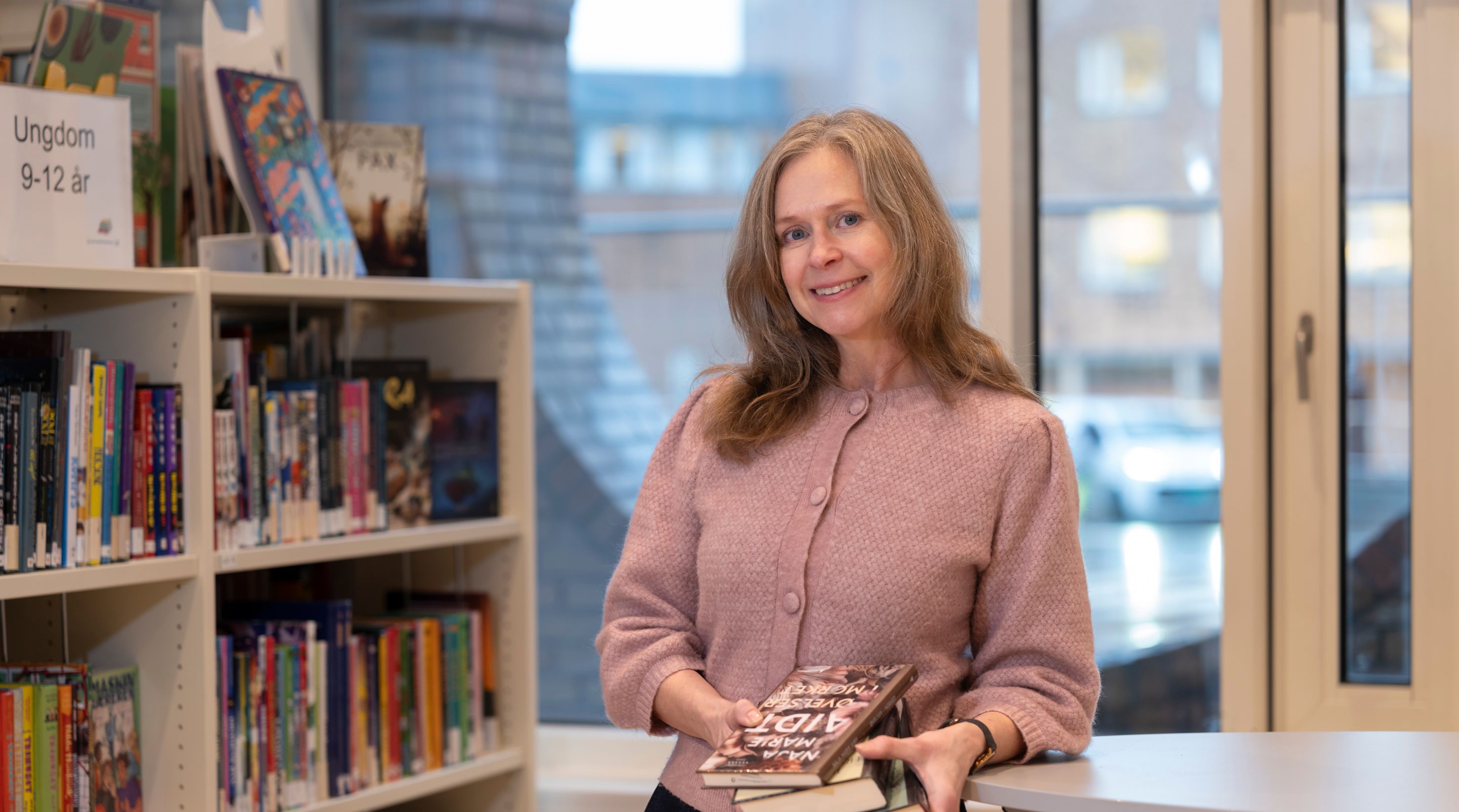 A smiling woman holds a stack of books in a library.