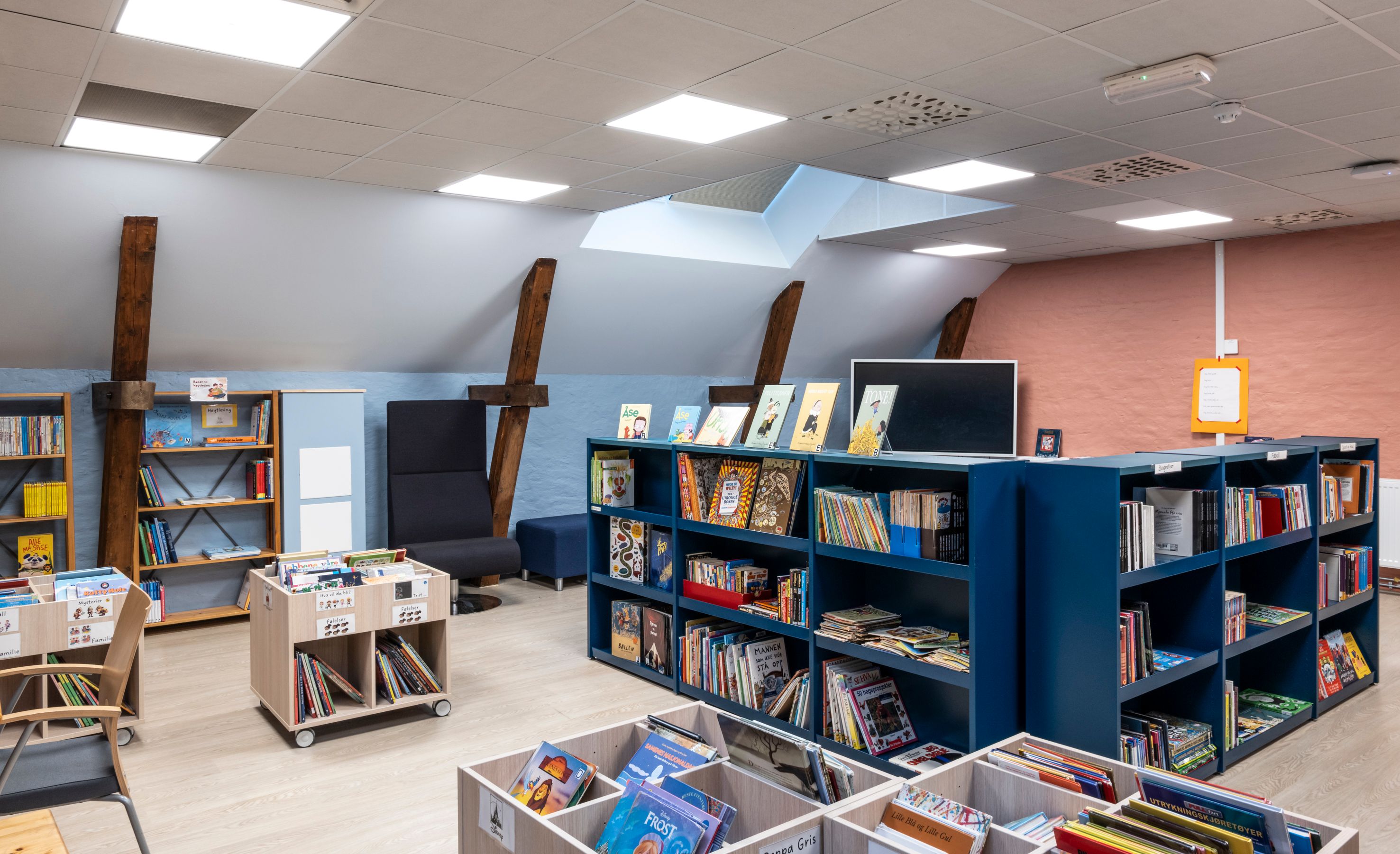 A brightly lit attic library with blue and wooden bookshelves filled with books, a large black chair, and exposed beams.