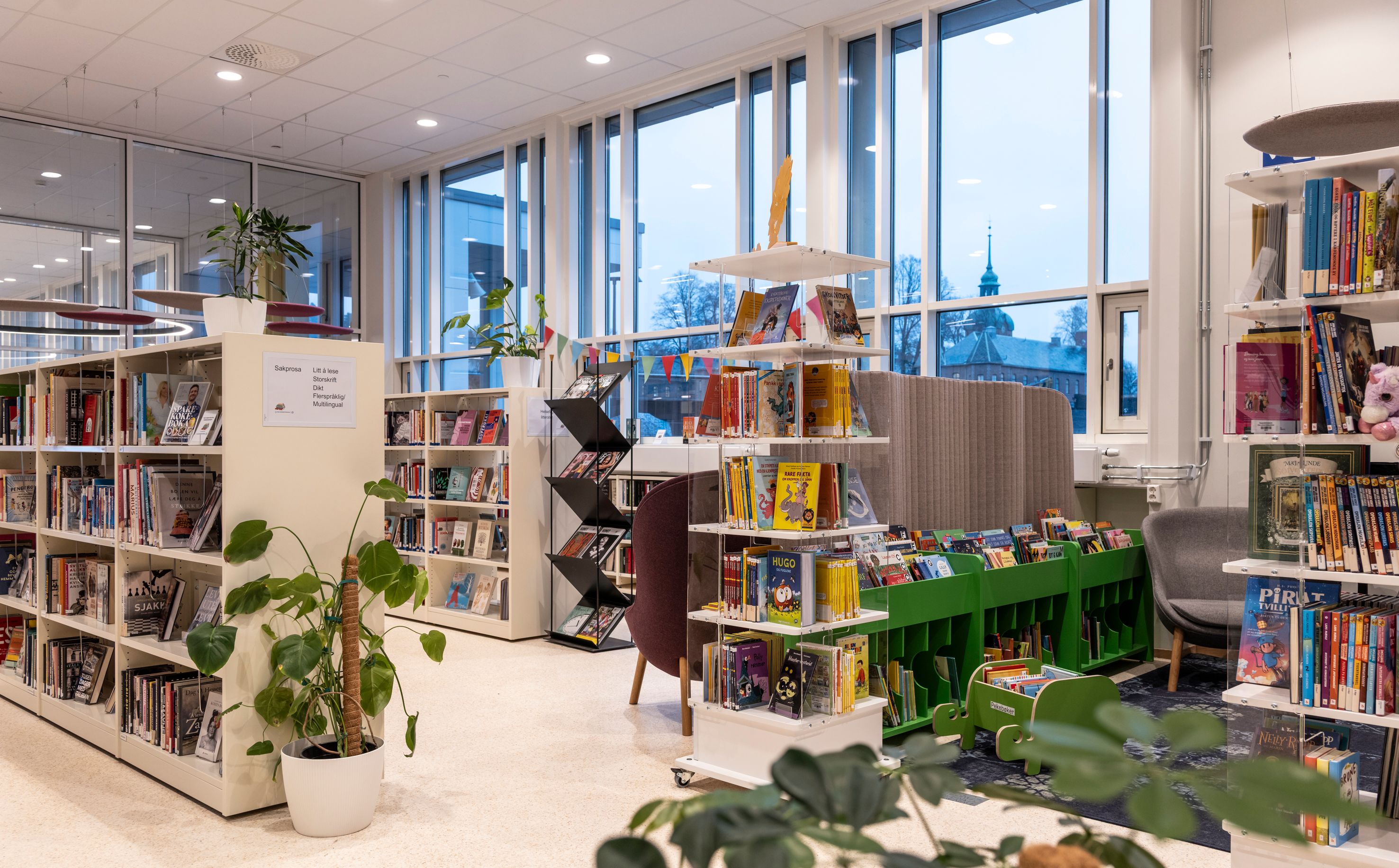 A bright, modern library interior with various bookshelves, green children's book displays, large windows, and potted plants.