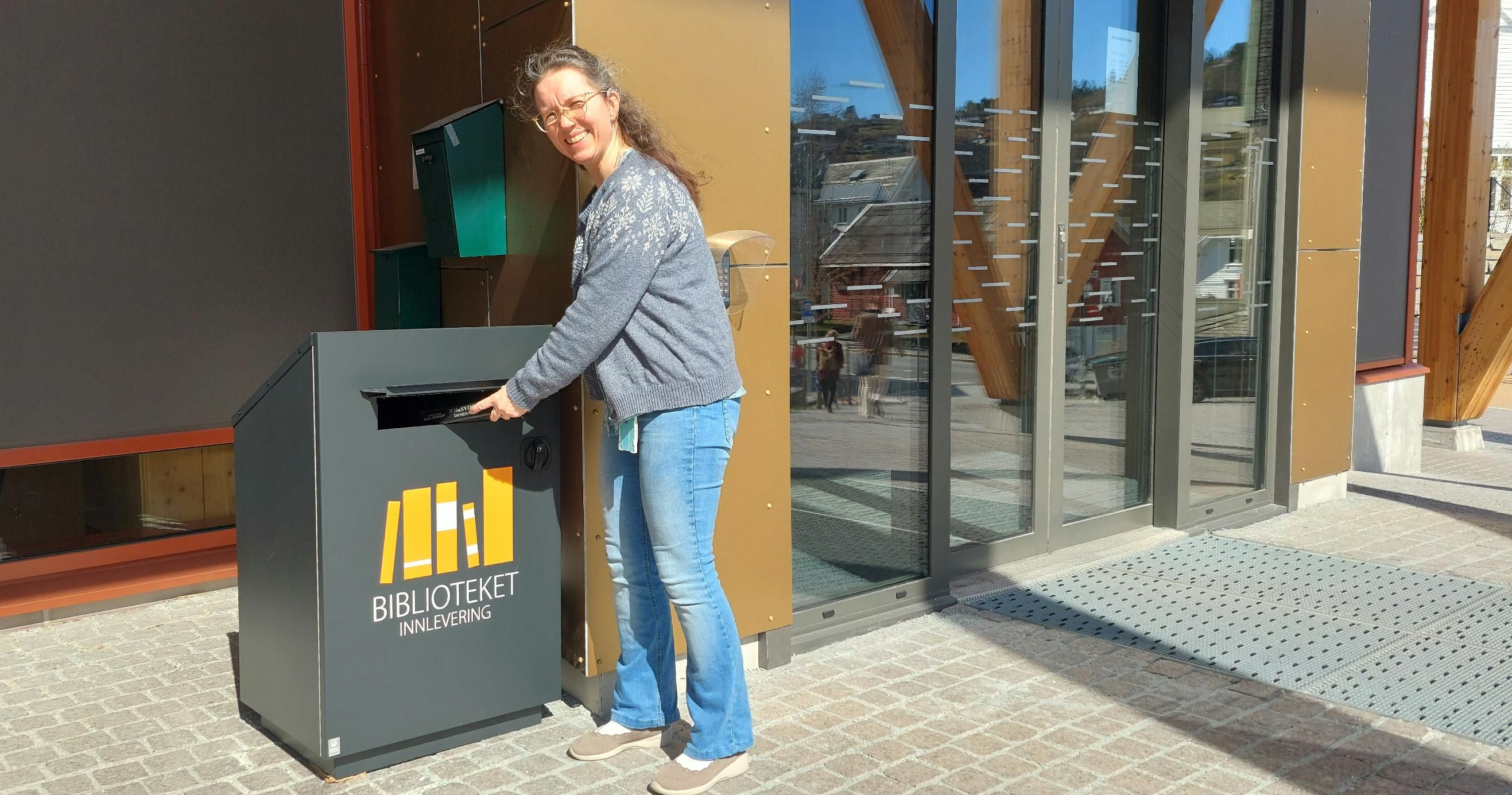 A smiling woman returns a book to an outdoor library drop box.