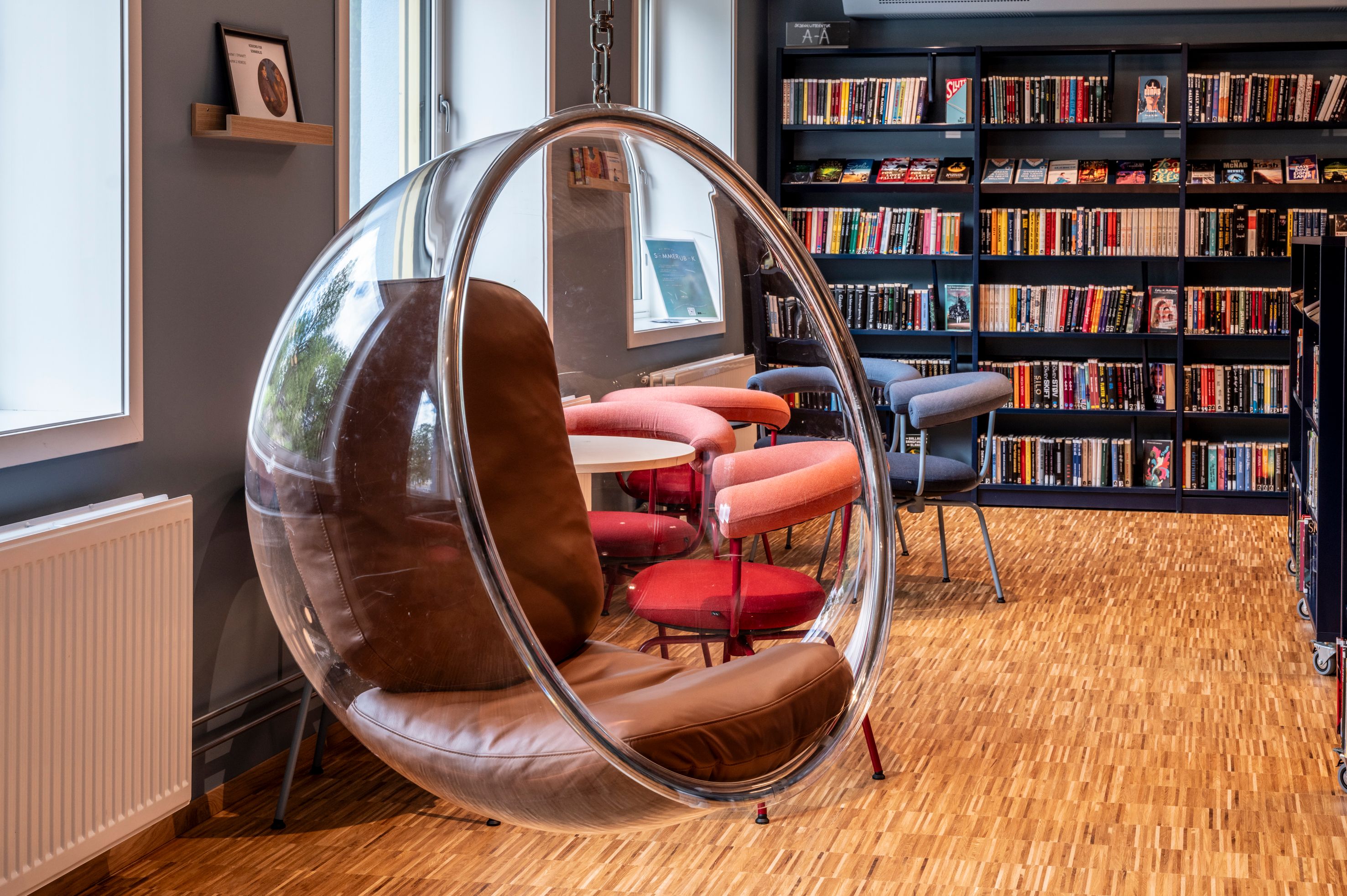 A clear bubble hanging chair with a brown cushion in a library, surrounded by colorful chairs, a table, and bookshelves.