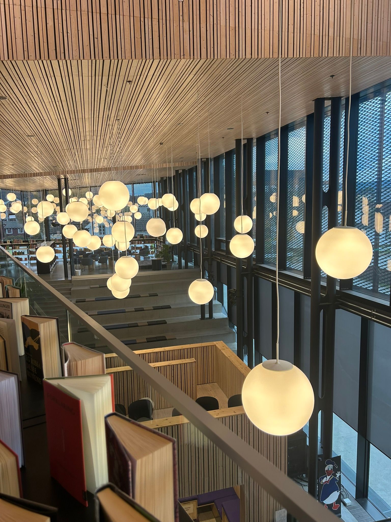 A bright, modern library interior with spherical pendant lights hanging from a wooden slatted ceiling, books along a railing, and large windows looking down to a lower level.