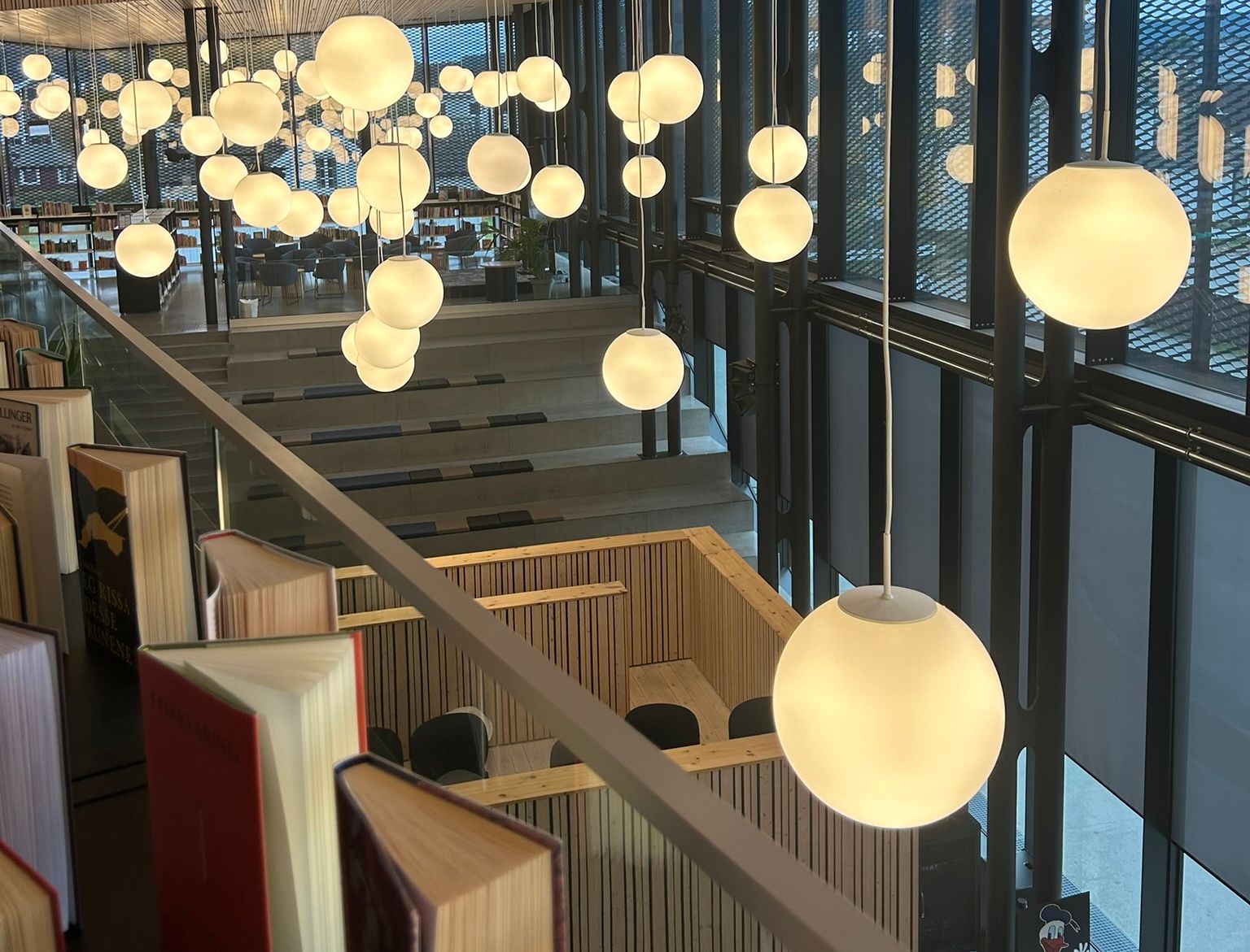 A bright, modern library interior with spherical pendant lights hanging from a wooden slatted ceiling, books along a railing, and large windows looking down to a lower level.