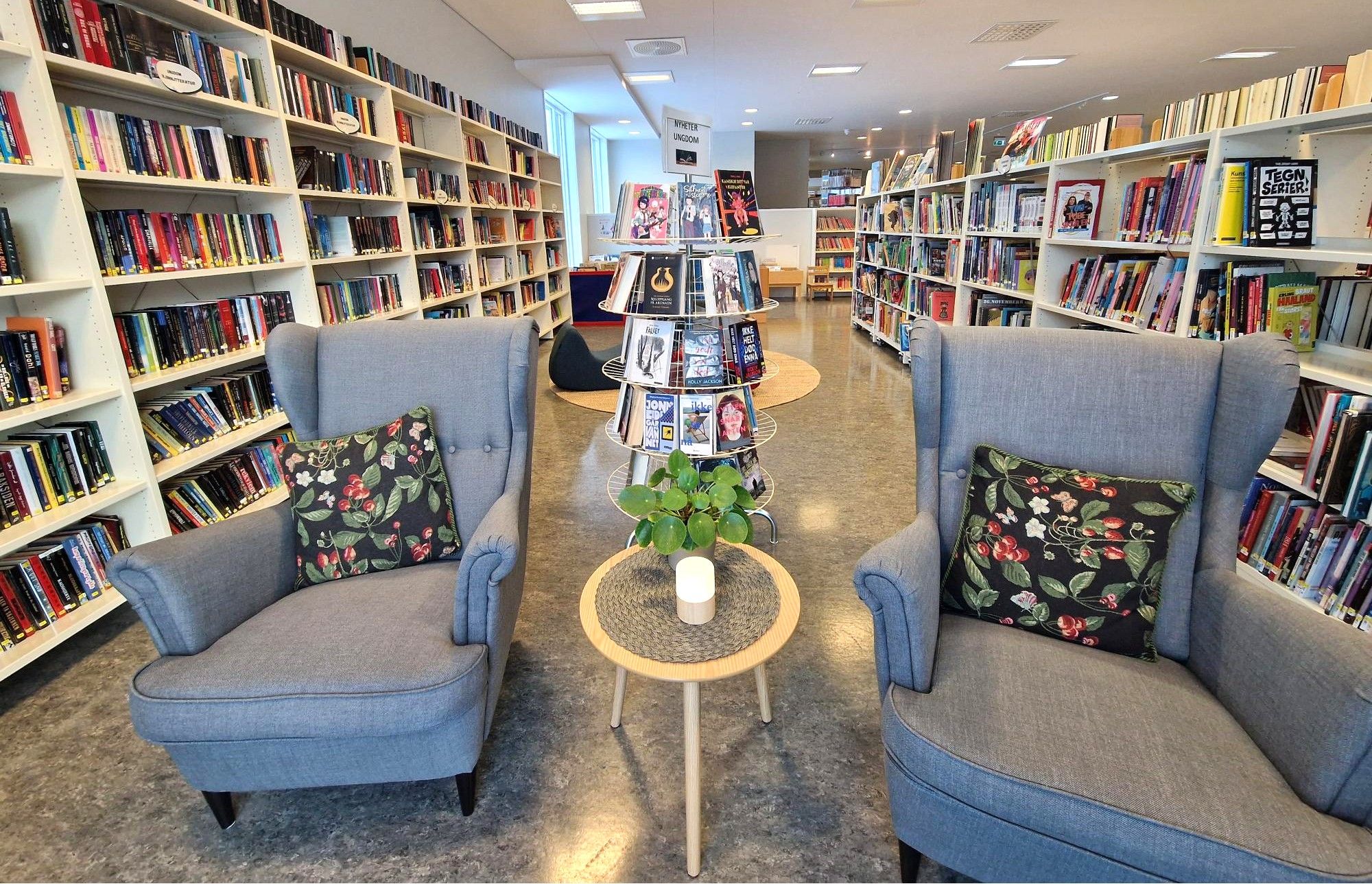 Two grey armchairs with floral pillows flank a small table in a modern library with rows of bookshelves.