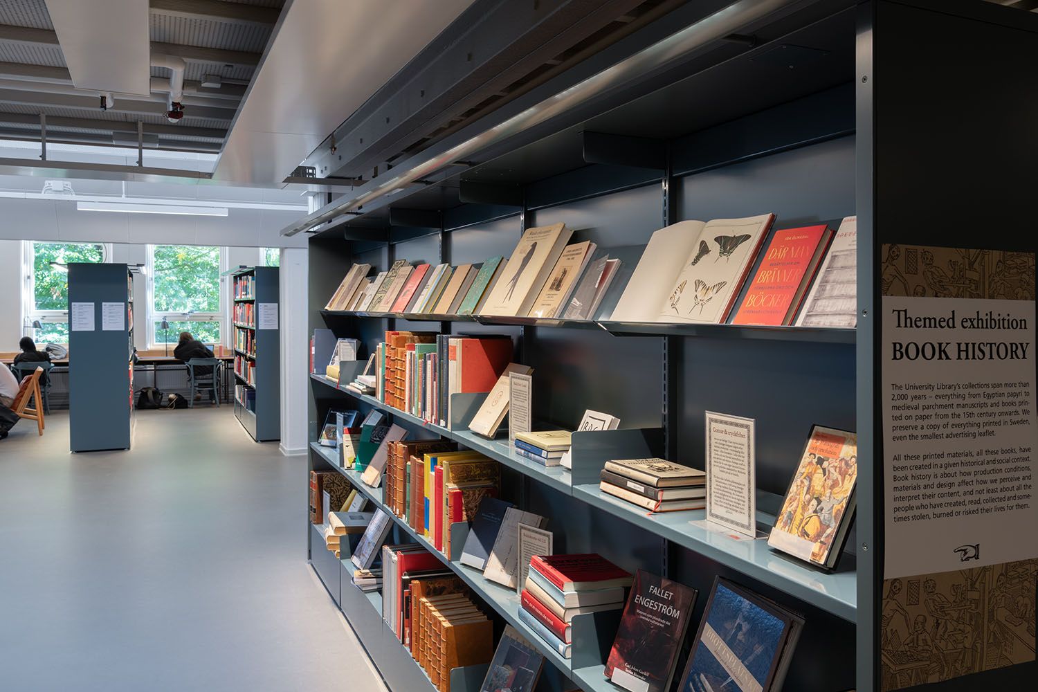 A modern library features a "Book History" exhibition on teal shelves, with people studying at tables in the background.